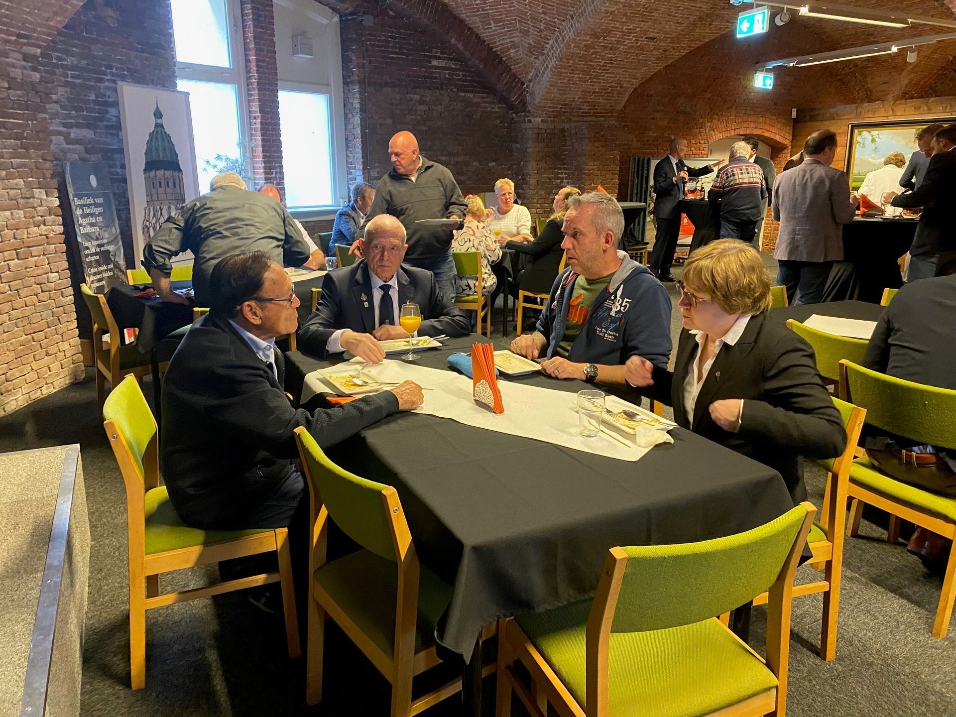 Mensen zitten aan tafels met zwarte tafelkleden in een kamer met gewelfde bakstenen muren en felgroene stoelen.