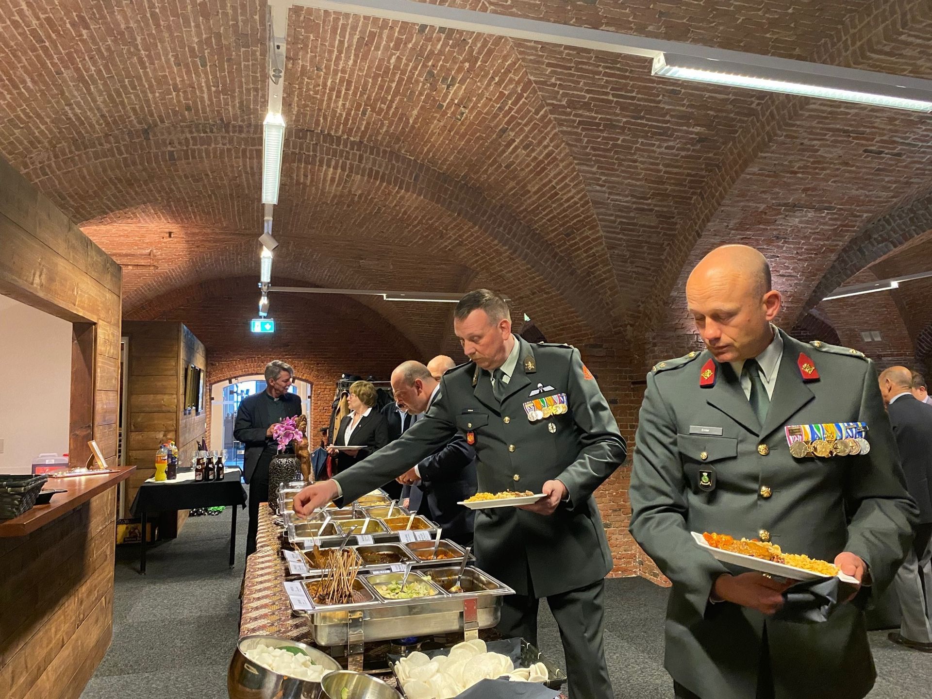 Officieren in formele militaire uniformen scheppen zelf hun eten op bij een buffet in een rustieke, bakstenen evenementenhal met gewelfd plafond.