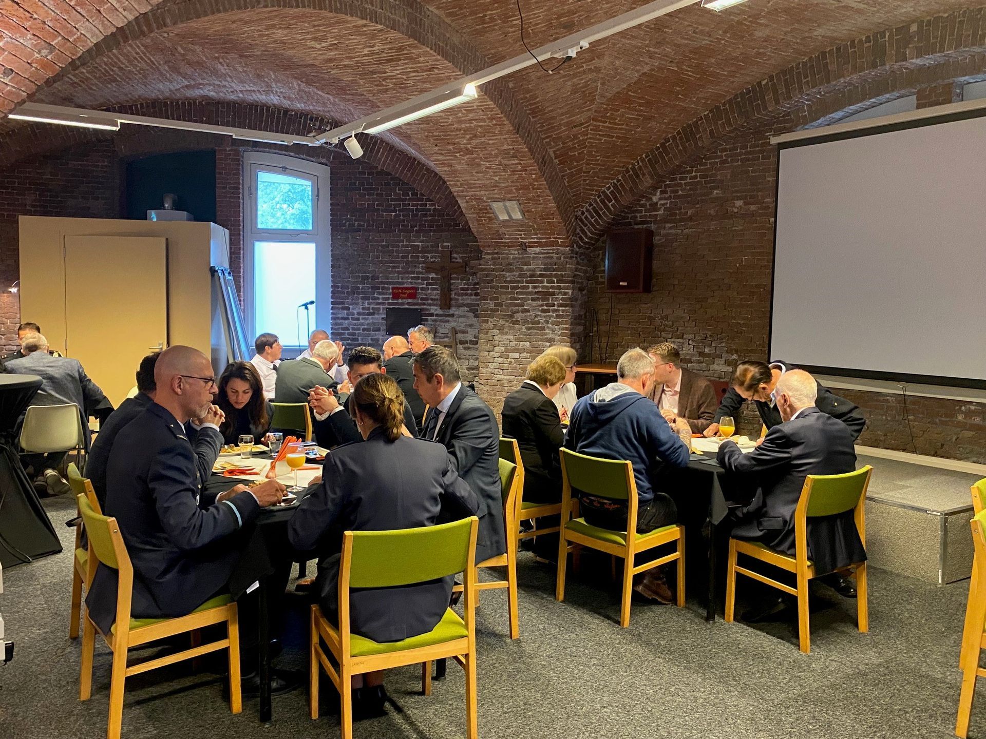 Mensen zitten aan tafels in een ruimte met een bakstenen gewelfd plafond en een projectiescherm, tijdens een vergadering of sociale bijeenkomst.