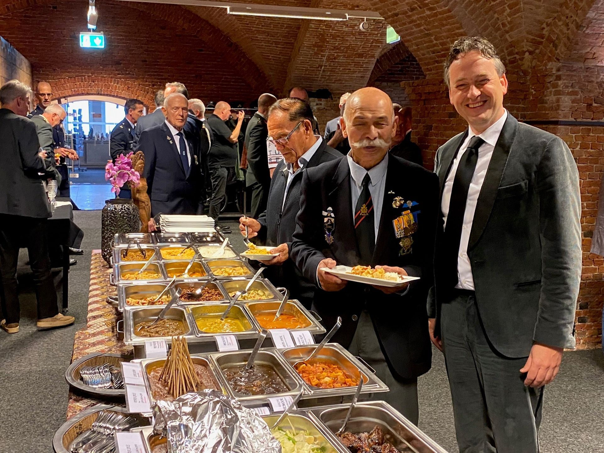 Mensen in pakken verzamelen zich bij een buffet in een rustieke zaal met bakstenen muren, waar twee mannen glimlachend borden met eten vasthouden.