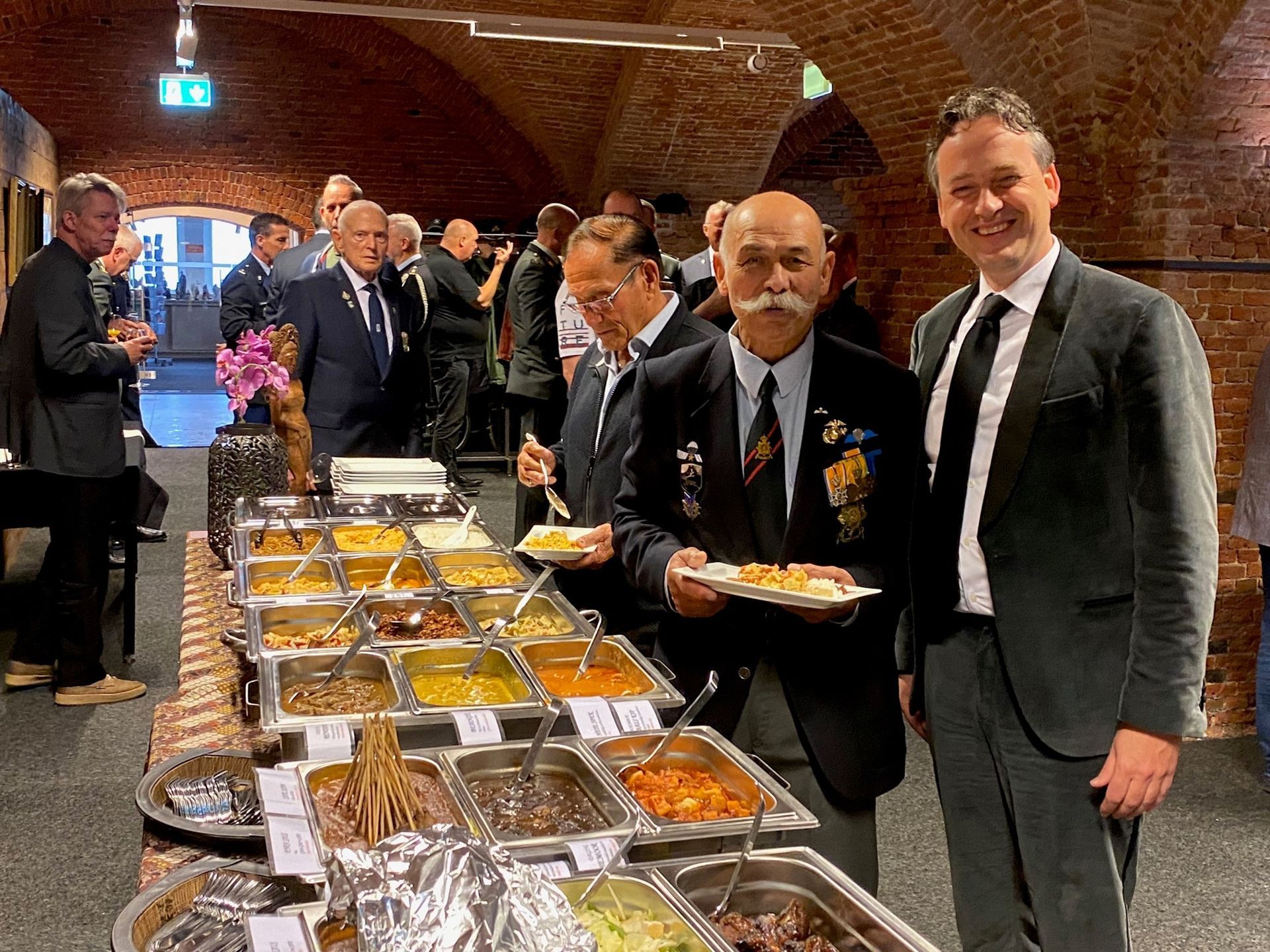 Mensen verzamelen zich bij een buffet in een ruimte met een gewelfd bakstenen plafond; twee mannen staan vooraan met borden vol eten.