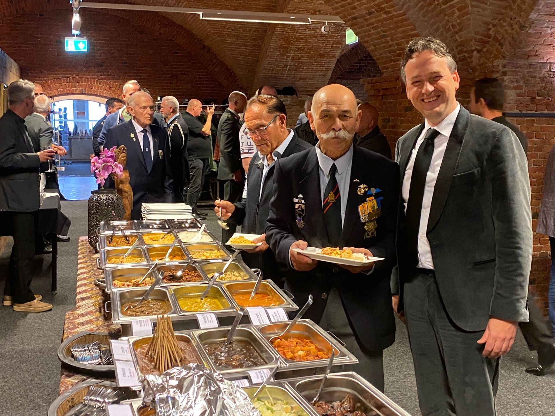 Mensen staan bij een buffet in een hal met bakstenen muren; een man in een blazer houdt een bord vast terwijl hij naar de camera lacht.