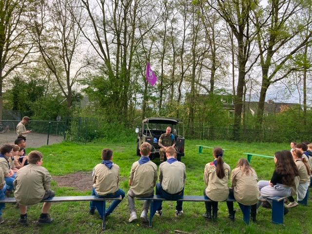 Een groep scouts in beige uniformen zit buiten op bankjes en luistert naar een spreker voor een donkergroen voertuig.