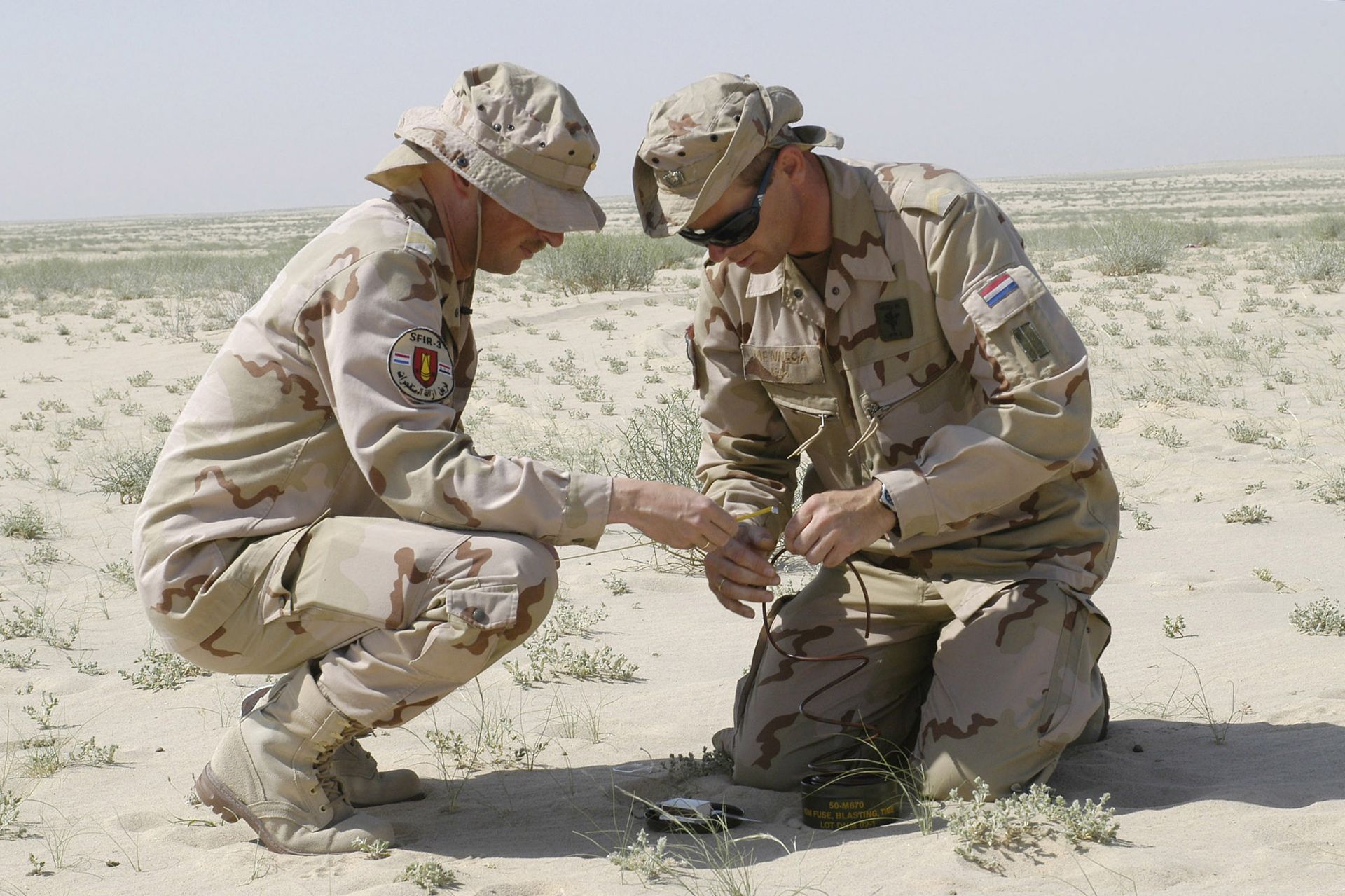 Twee soldaten in woestijncamouflage-uniformen knielen in het zand en onderzoeken een klein voorwerp op de grond.