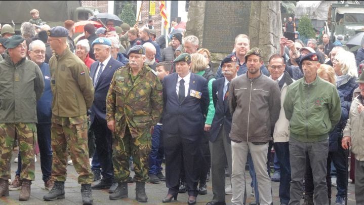 Een diverse groep veteranen, in militaire en burgerkleding, staat in een plechtige formatie in de openlucht tijdens een herdenkingsdienst.