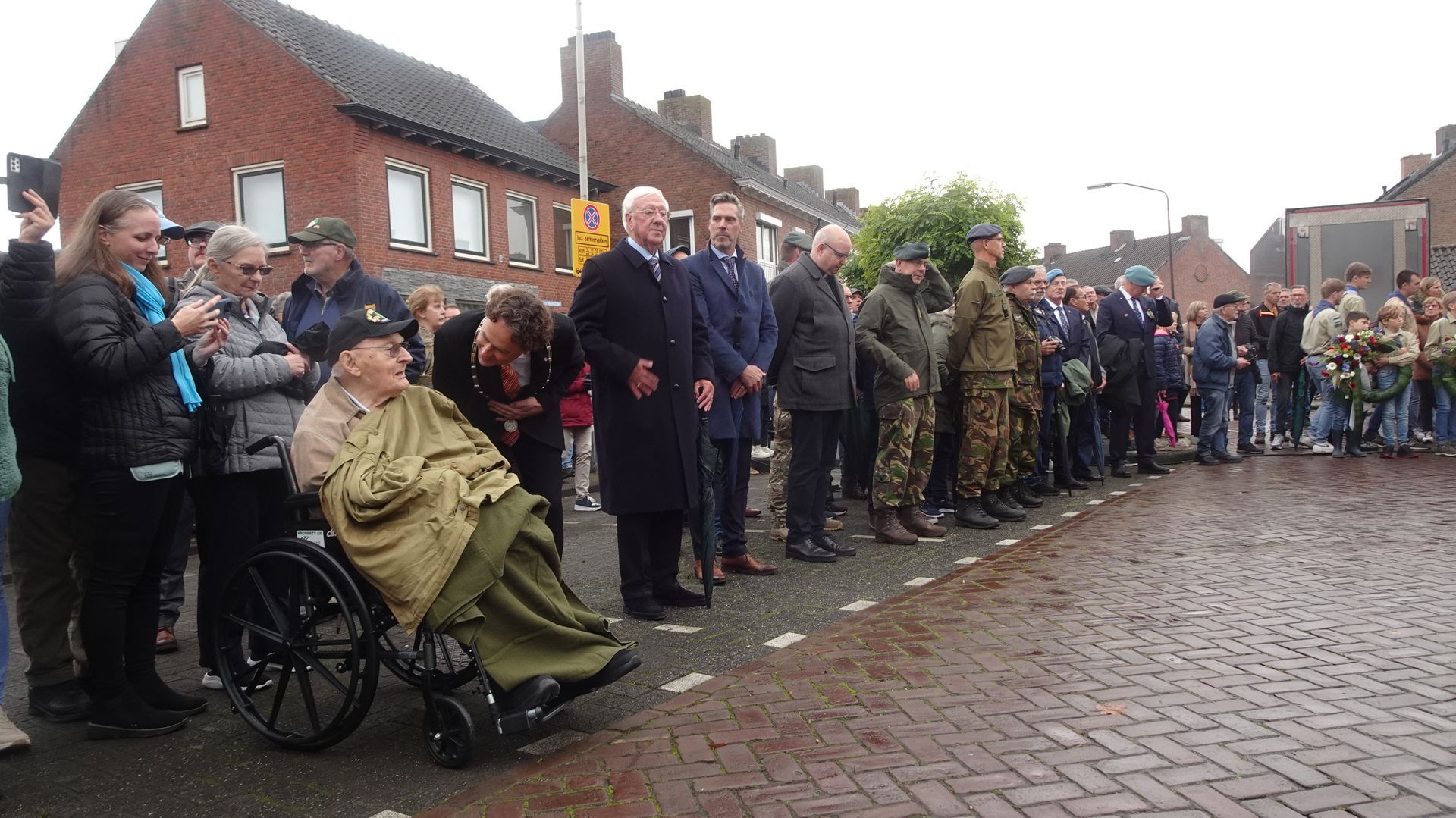Mensen staan ​​in een rij op een bakstenen straat tijdens een ceremonie in de openlucht, waaronder een persoon in een rolstoel.