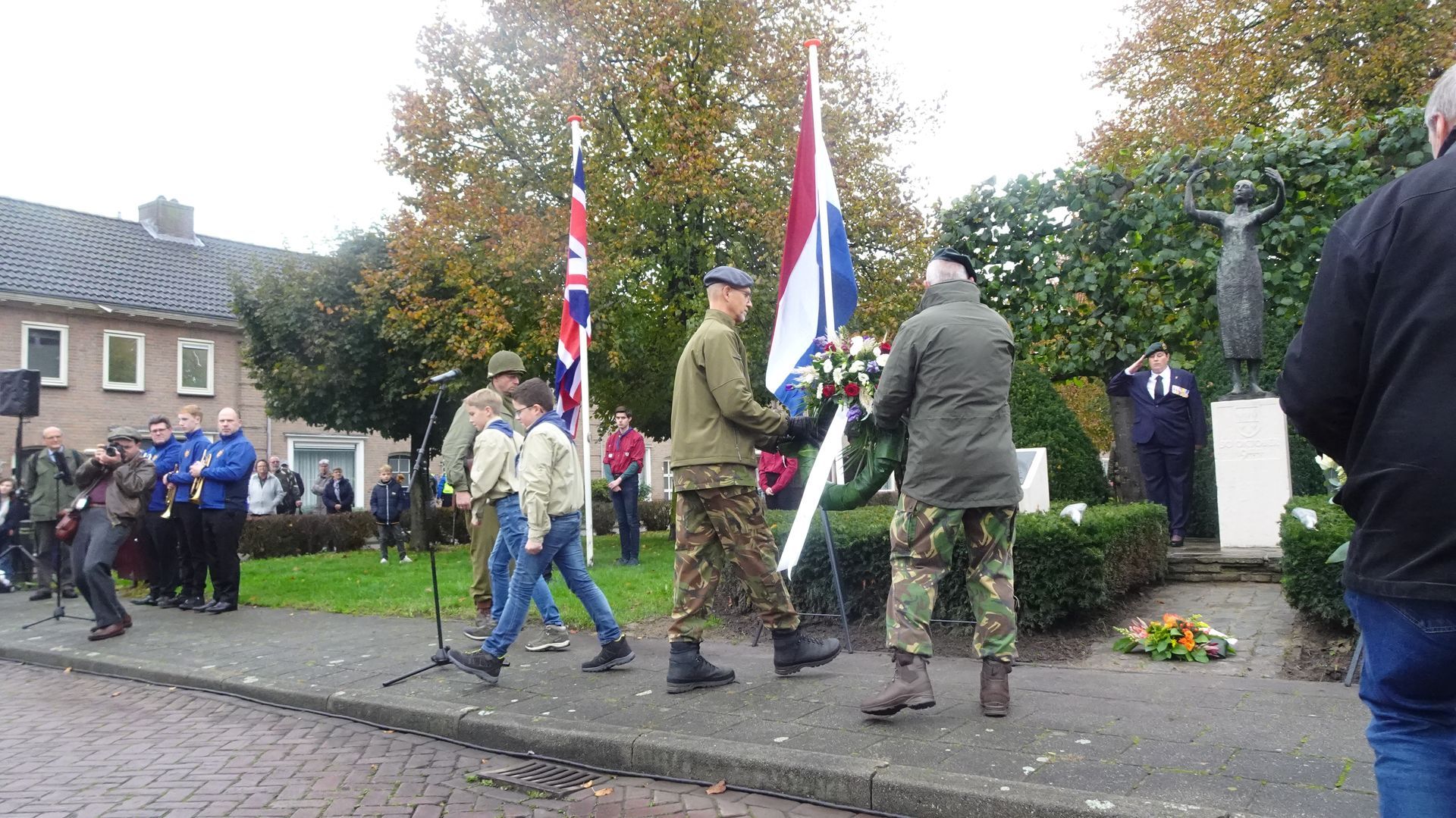 Mensen in militaire uniformen en vrijetijdskleding leggen een krans bij een monument tijdens een herdenkingsceremonie in de openlucht.