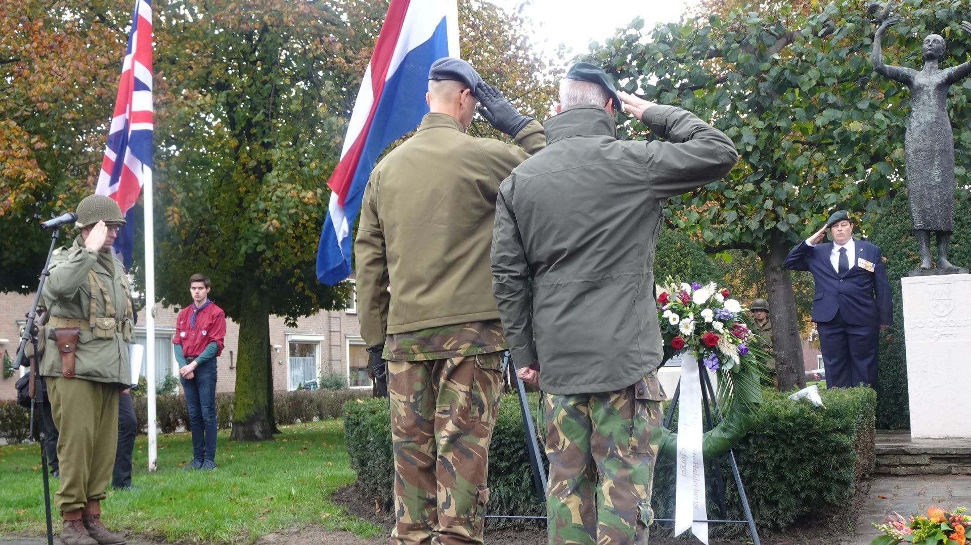 Mensen in militaire en burgerkleding staan ​​en brengen een saluut bij een gedenkplaats, met vlaggen en een standbeeld op de achtergrond.