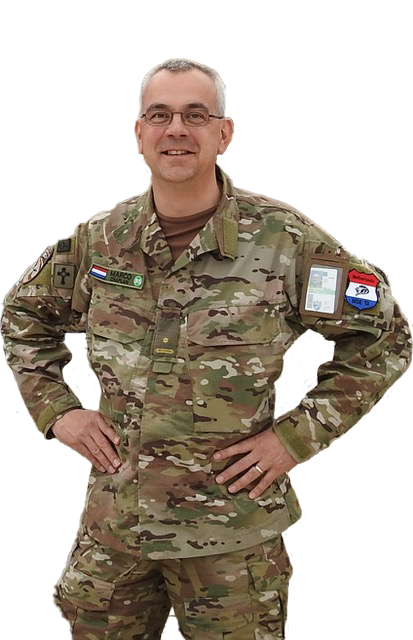 Een soldaat in camouflage-uniform met bril en een vriendelijke uitdrukking staat met zijn handen in zijn zij tegen een witte achtergrond.
