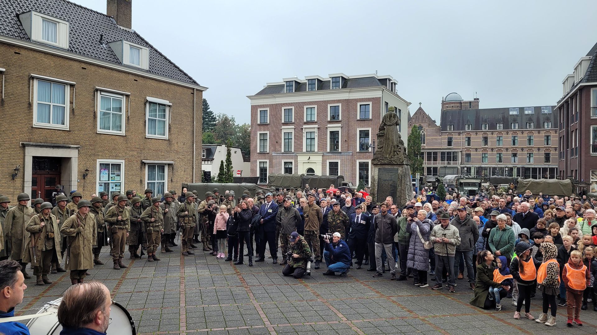 Een grote, diverse menigte, waaronder soldaten in uniform, verzamelt zich op een geplaveid stadsplein om een ceremonie in de openlucht bij te wonen.