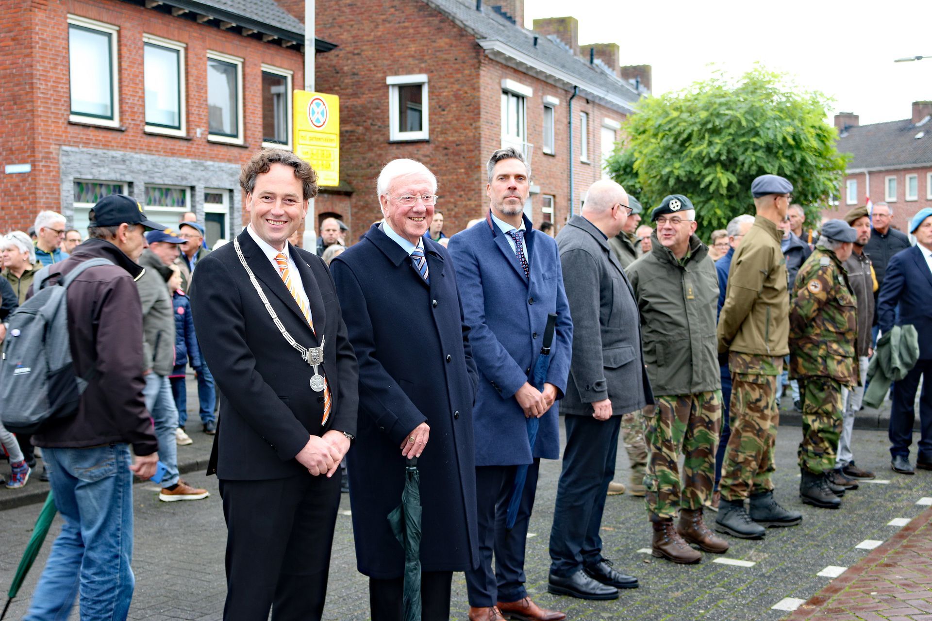 Een groep mensen in formele en militaire kleding staat in een rij buiten om een herdenkingsplechtigheid bij te wonen.