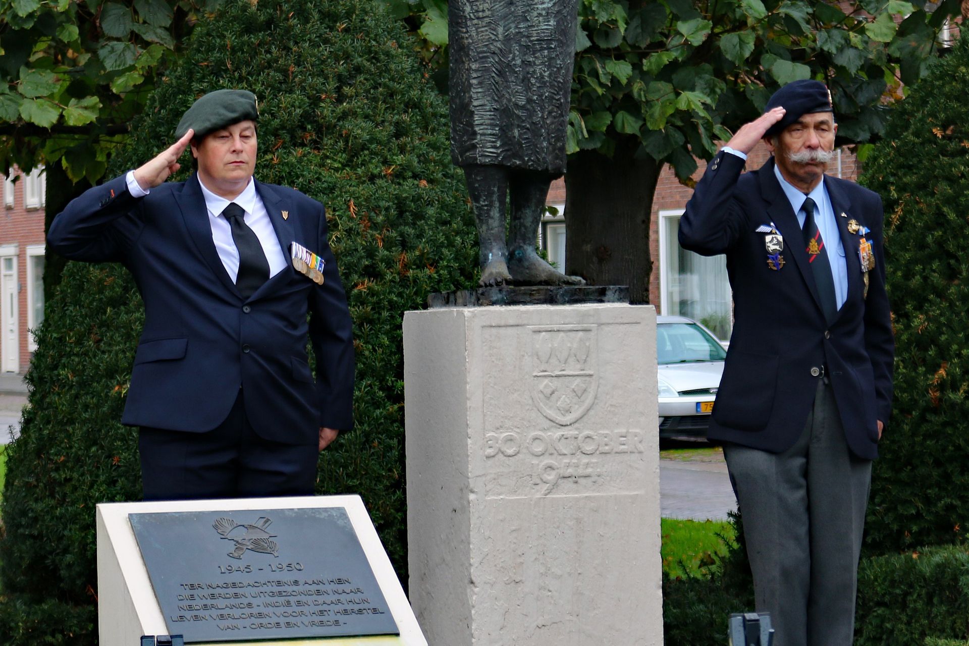 Twee personen in colberts en baretten staan in de houding en brengen een saluut voor een stenen monument en standbeeld in een openluchtomgeving.