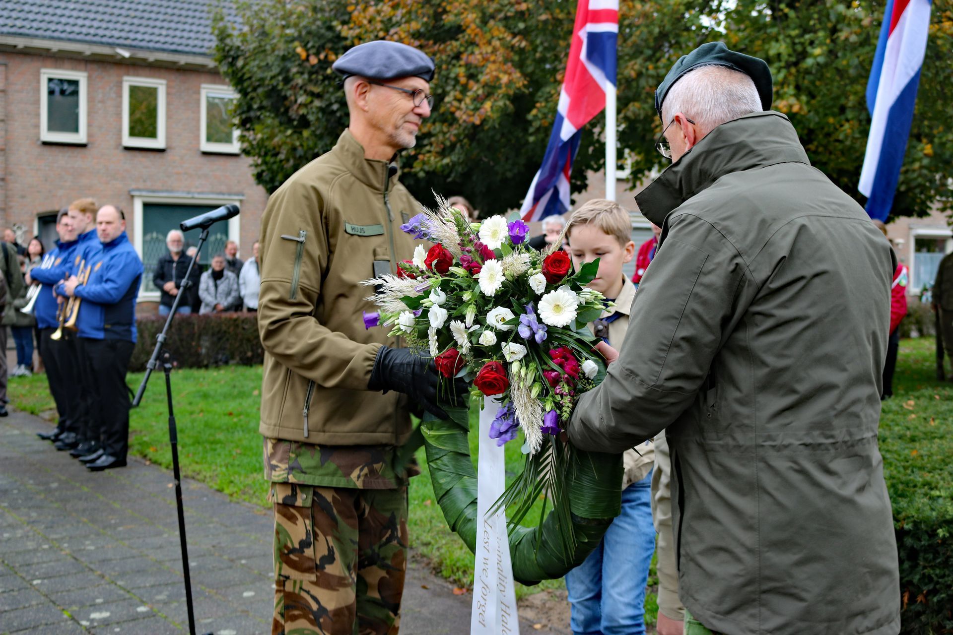 Twee mensen in outdoorkleding houden een grote, kleurrijke rouwkrans vast tijdens een openluchtceremonie voor vlaggen.