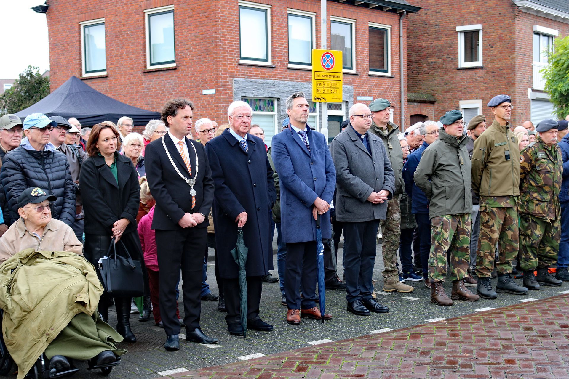 Een diverse groep mensen in formele en militaire kleding staat buiten tijdens een herdenkingsceremonie in de openlucht.