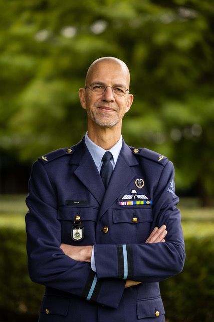 Een militair officier in een blauw uniform met medailles, met de armen over elkaar, staat buiten tegen een wazige groene achtergrond.