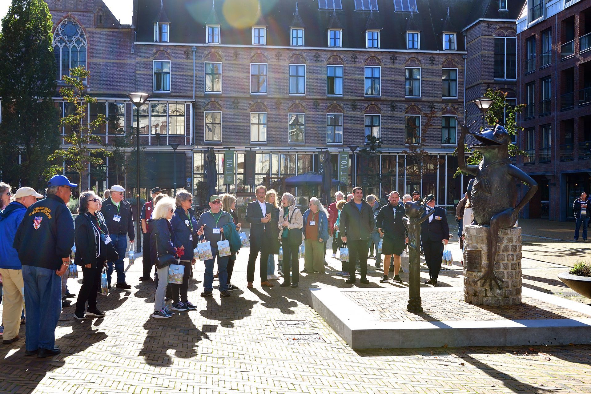 Een groep mensen staat op een bakstenen plein voor een bronzen beeld van een figuur met grote voeten, vlakbij historische gebouwen.