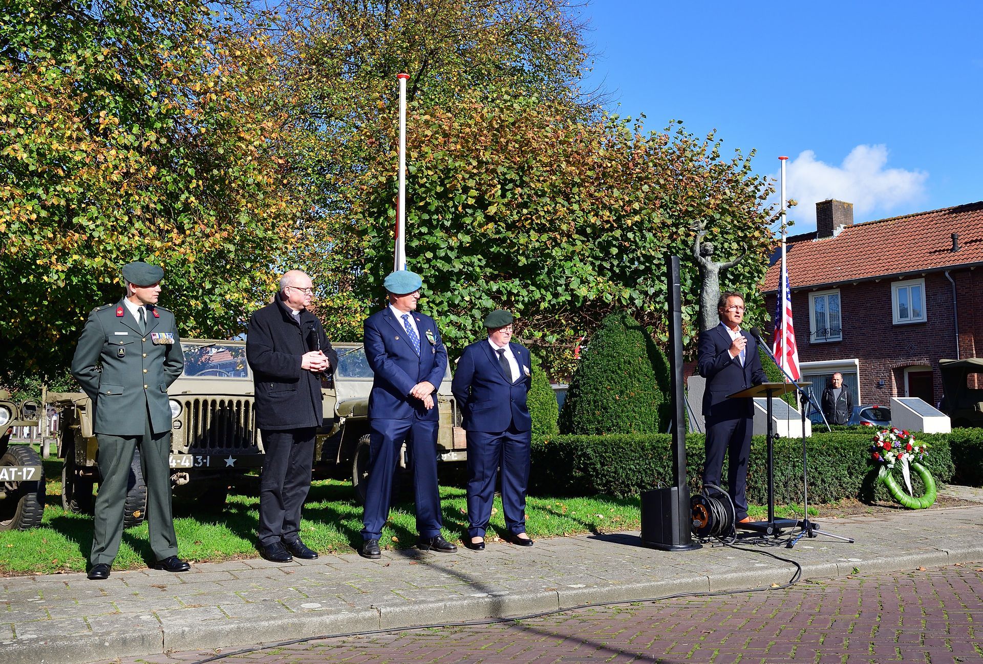 Vier personen, waaronder drie in uniform, staan ​​buiten tijdens een ceremonie in de buurt van een voertuig, bomen en een bakstenen gebouw.