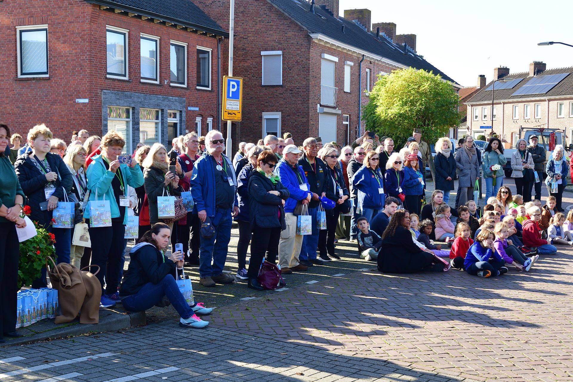 Een menigte mensen staat en zit op een geplaveide straat voor bakstenen gebouwen en kijkt naar een openbaar evenement in de openlucht.