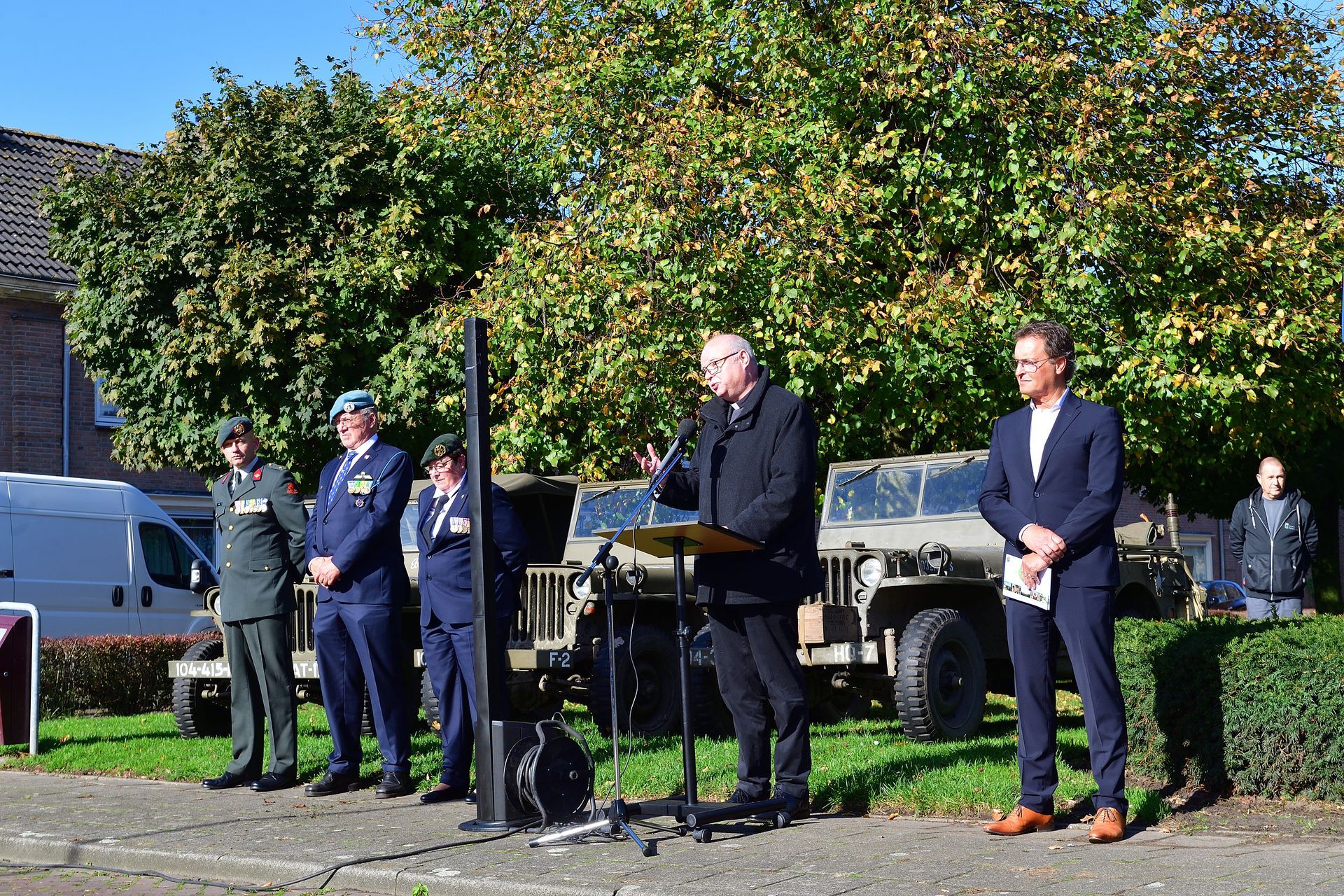 Een man spreekt vanaf een podium tijdens een herdenkingsdienst in de openlucht, met drie geüniformeerde militairen en een burger in de buurt.