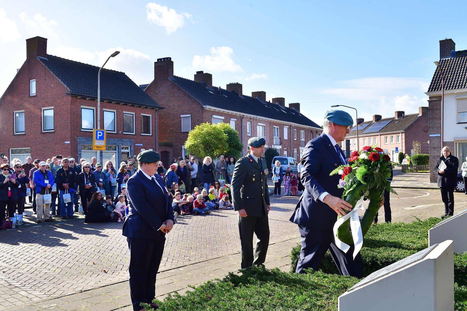Drie personen in militaire uniformen staan ​​voor een monument, waarvan er één een bloemenkrans neerlegt, terwijl een menigte toekijkt.