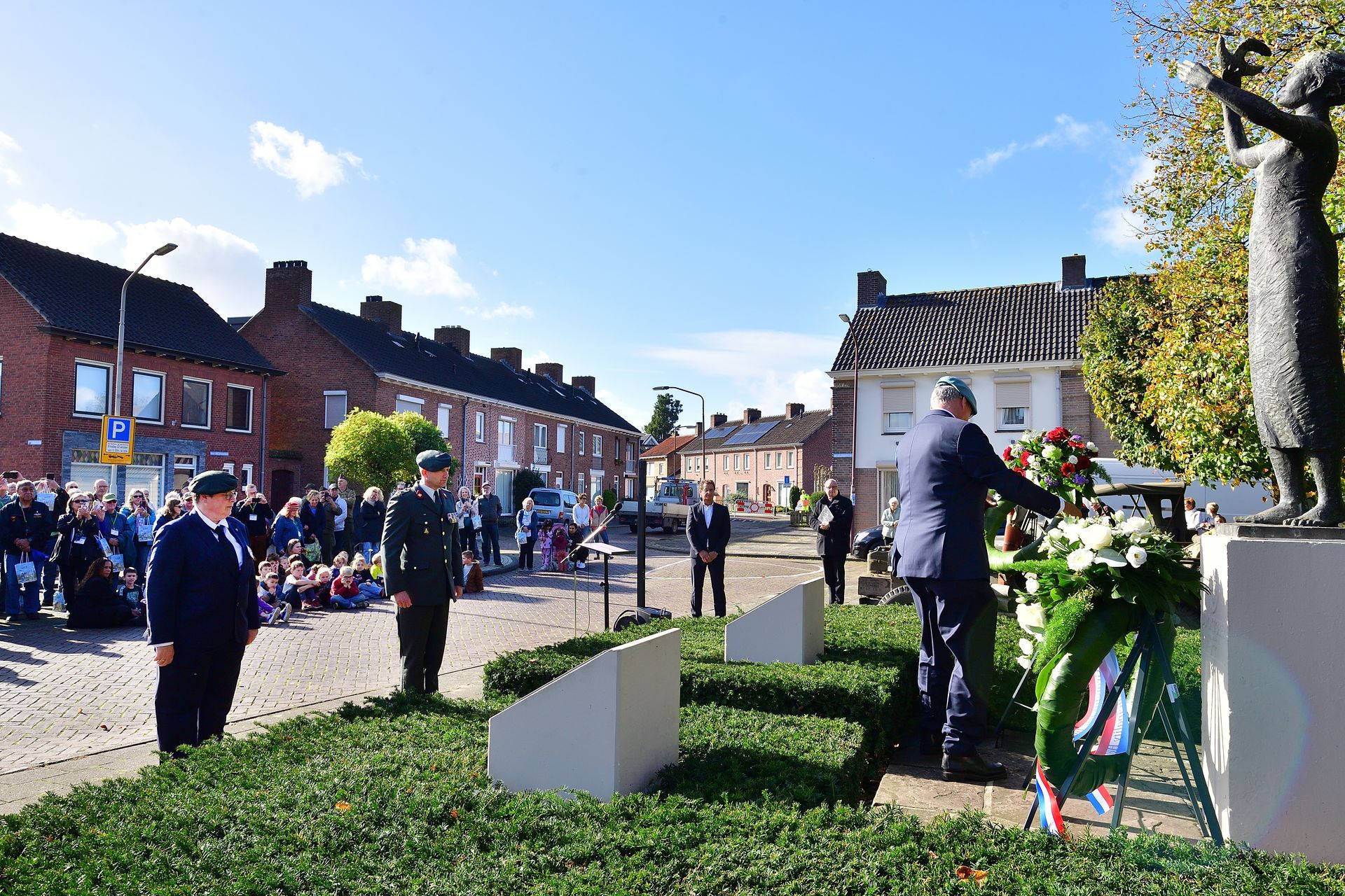 Tijdens een herdenkingsceremonie op een openbaar plein legt iemand een krans bij een monument, met toeschouwers op de achtergrond.