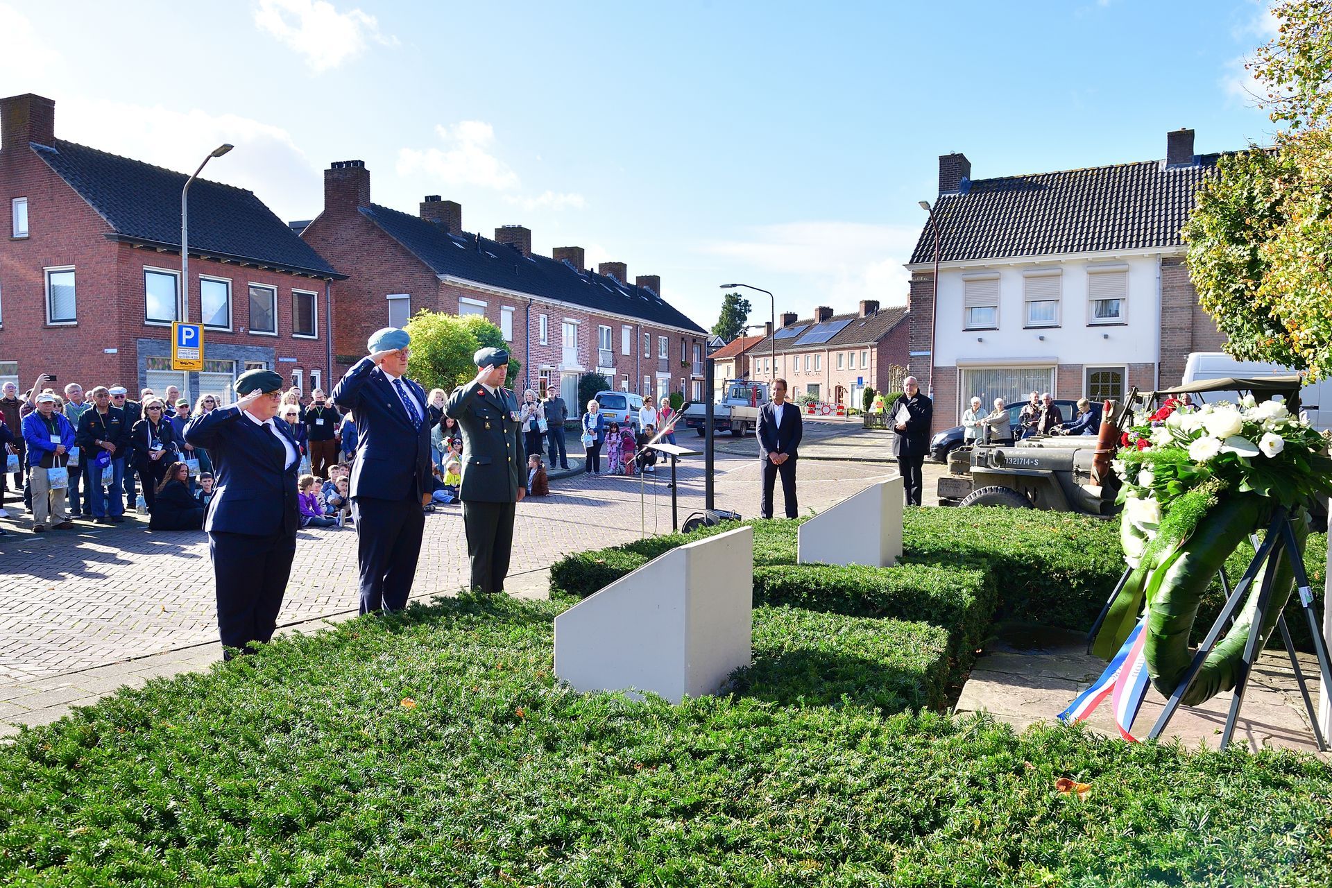 Drie militairen in uniform brengen een saluut bij een herdenkingsplek, omringd door een menigte en woongebouwen.