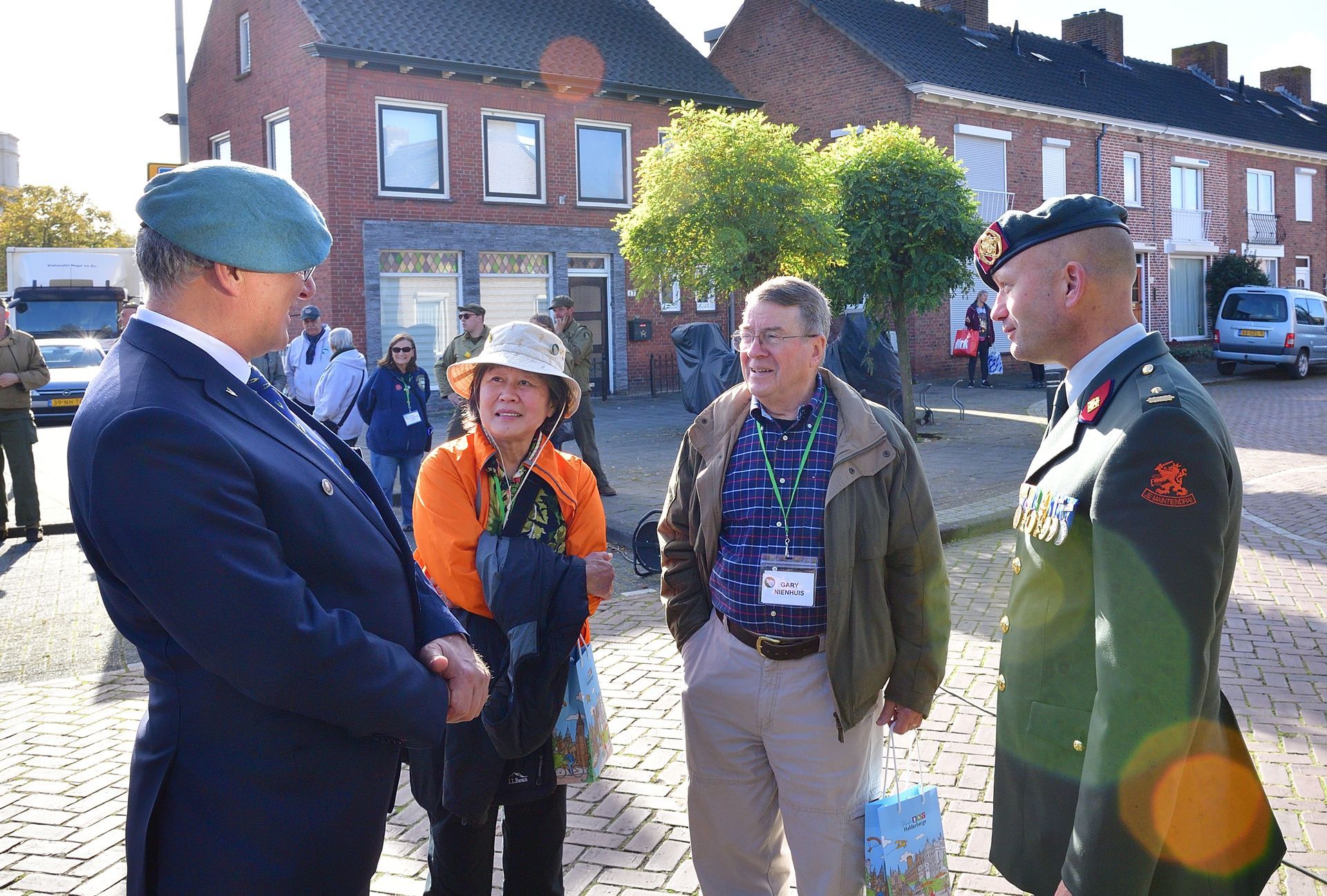Vier mensen, twee in militair uniform en twee in vrijetijdskleding, staan ​​buiten op een met bakstenen bestrate stadsplein.
