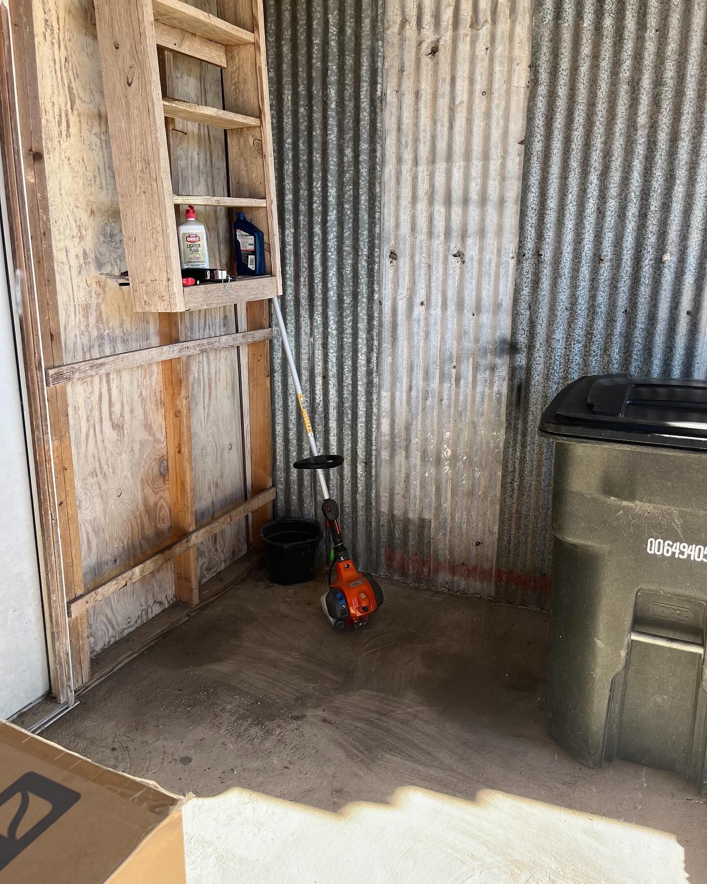 A weed trimmer stands in a shed with wooden wall shelves and a corrugated metal wall next to a large trash bin.