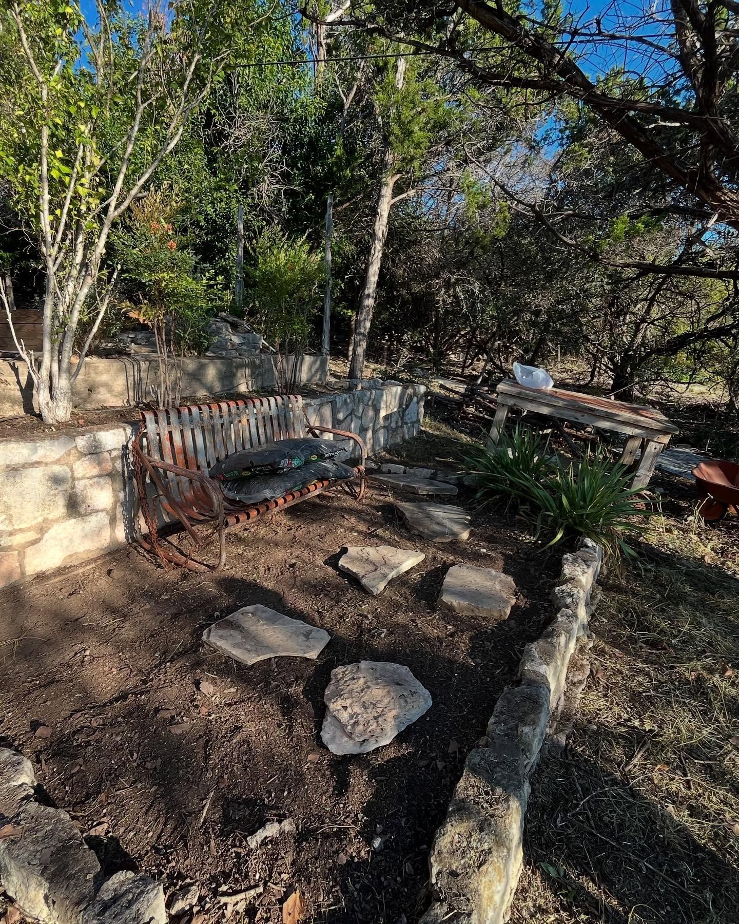 A rustic wooden bench sits in a sunlit garden beside a stone wall and path made of flat stepping stones.
