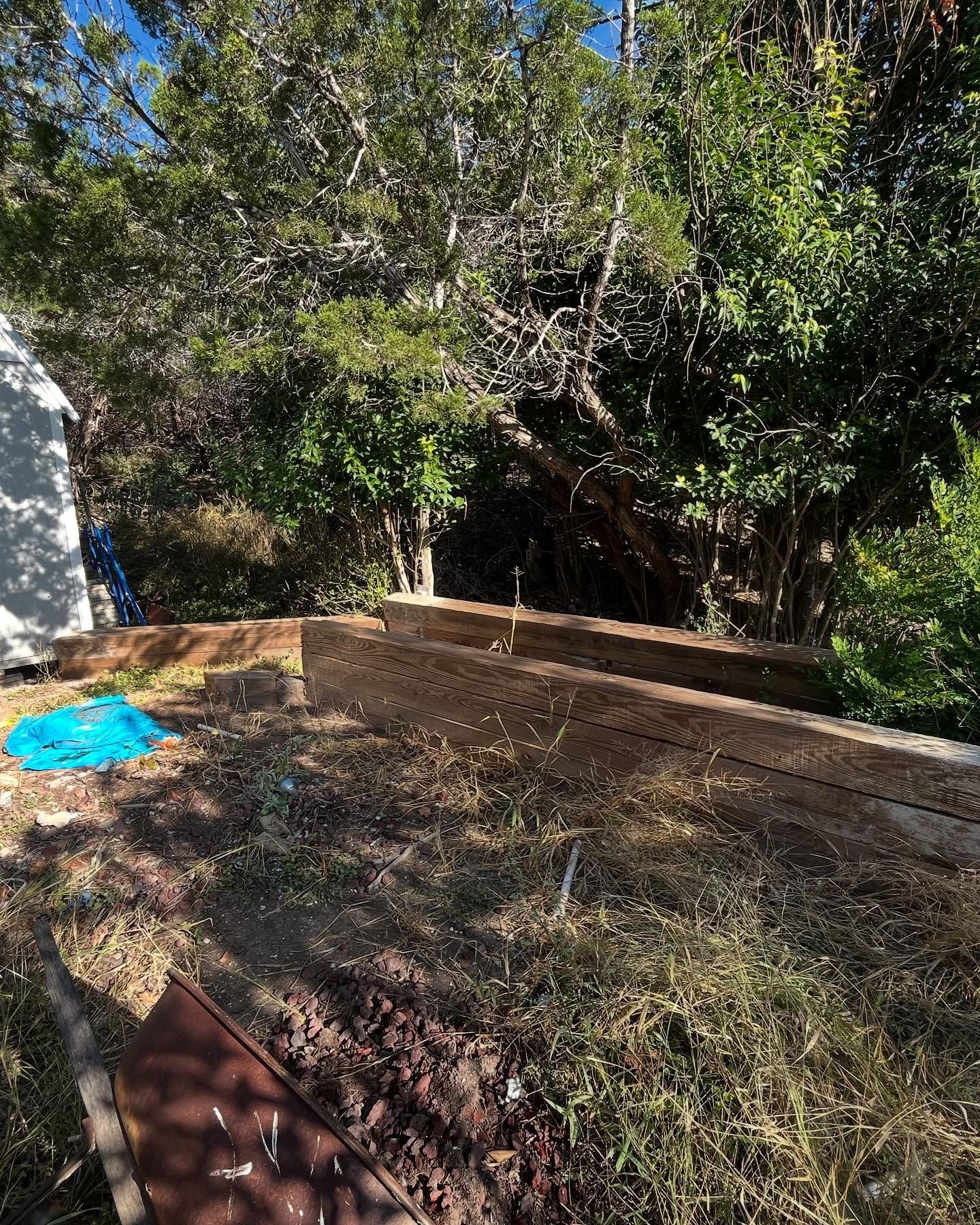 Stacked wooden retaining wall timbers sit in a backyard, partially obscured by trees and shadows under a clear blue sky.