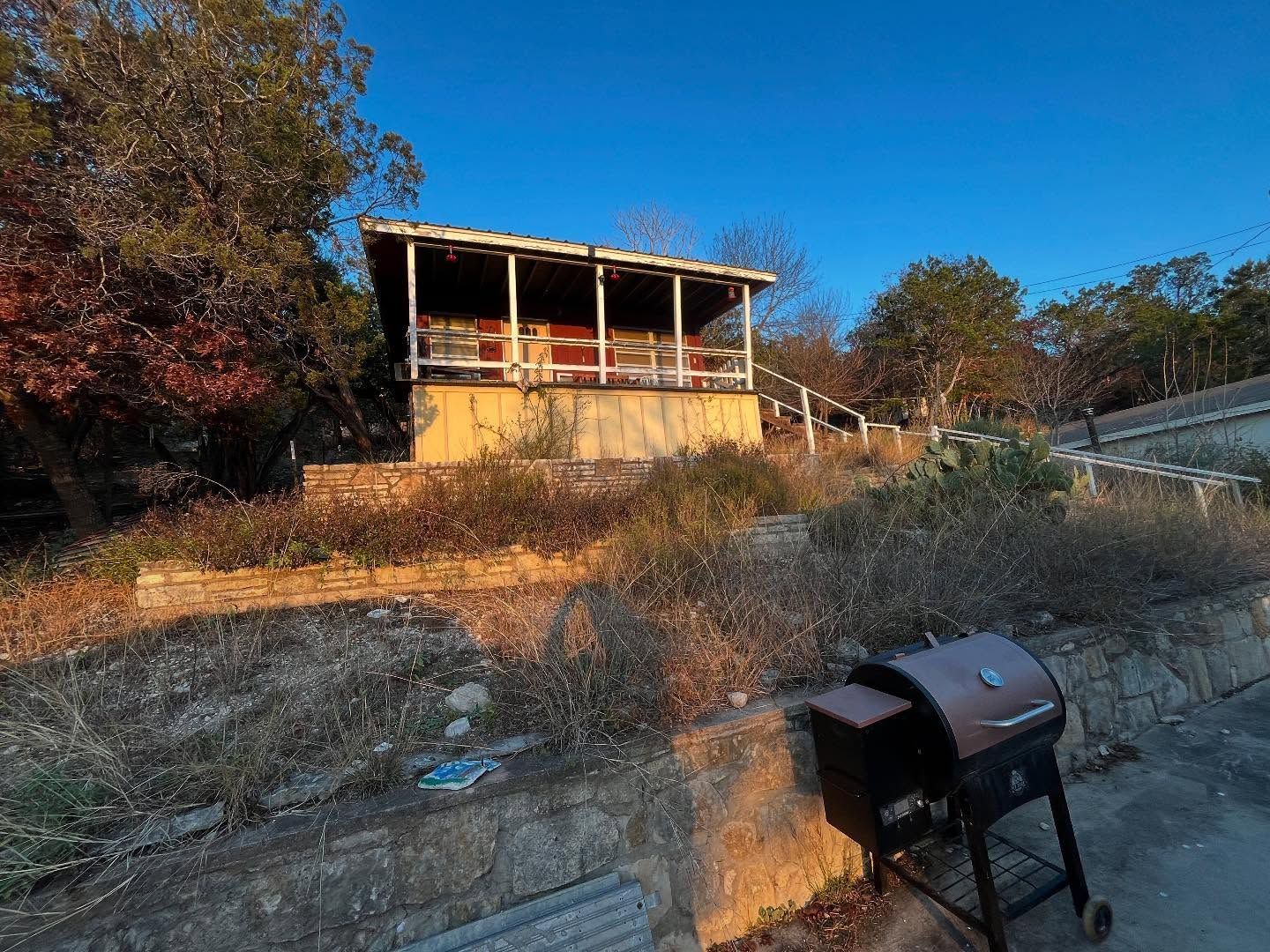 A tan cabin with a wooden porch sits on a raised stone landscape at sunset, with a charcoal grill in the foreground.