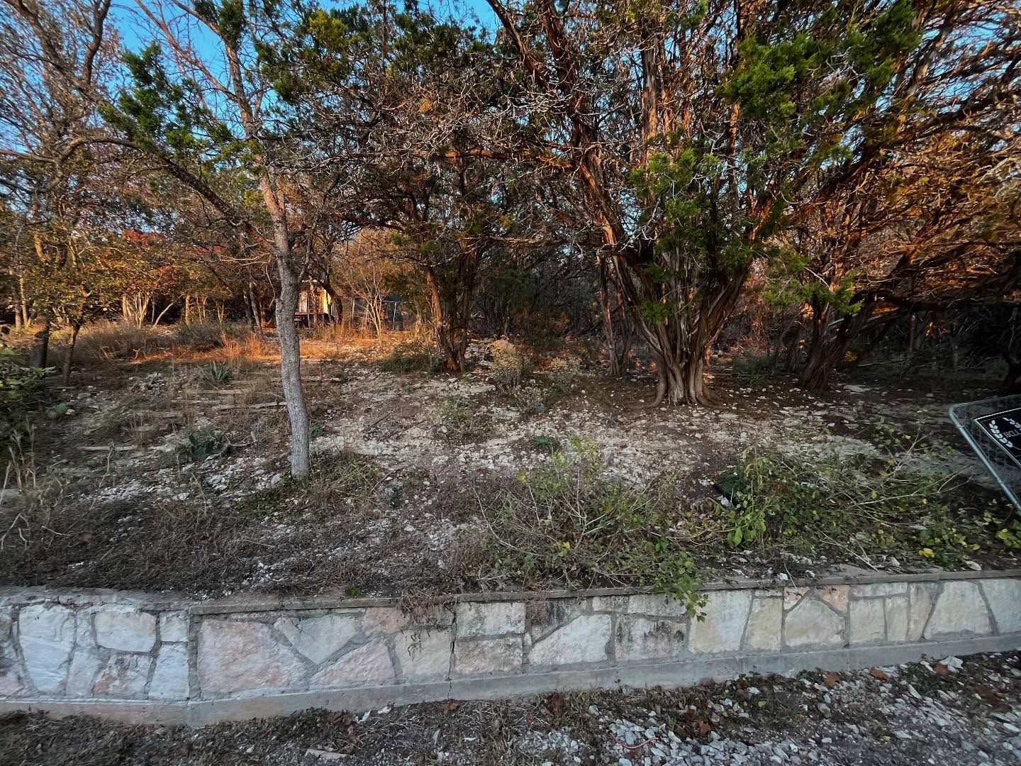 A stone retaining wall borders a sloped, rocky yard filled with scrub brush and several mature cedar trees at sunset.