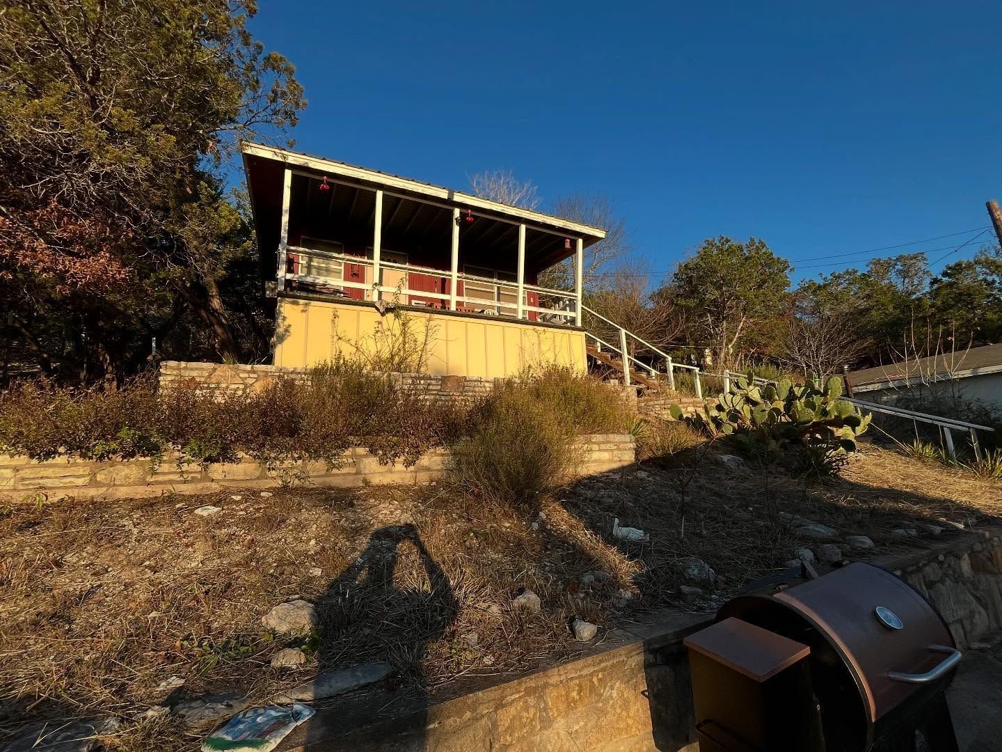 A elevated wooden cabin with a porch stands on a hillside, illuminated by warm sunlight against a clear blue sky.
