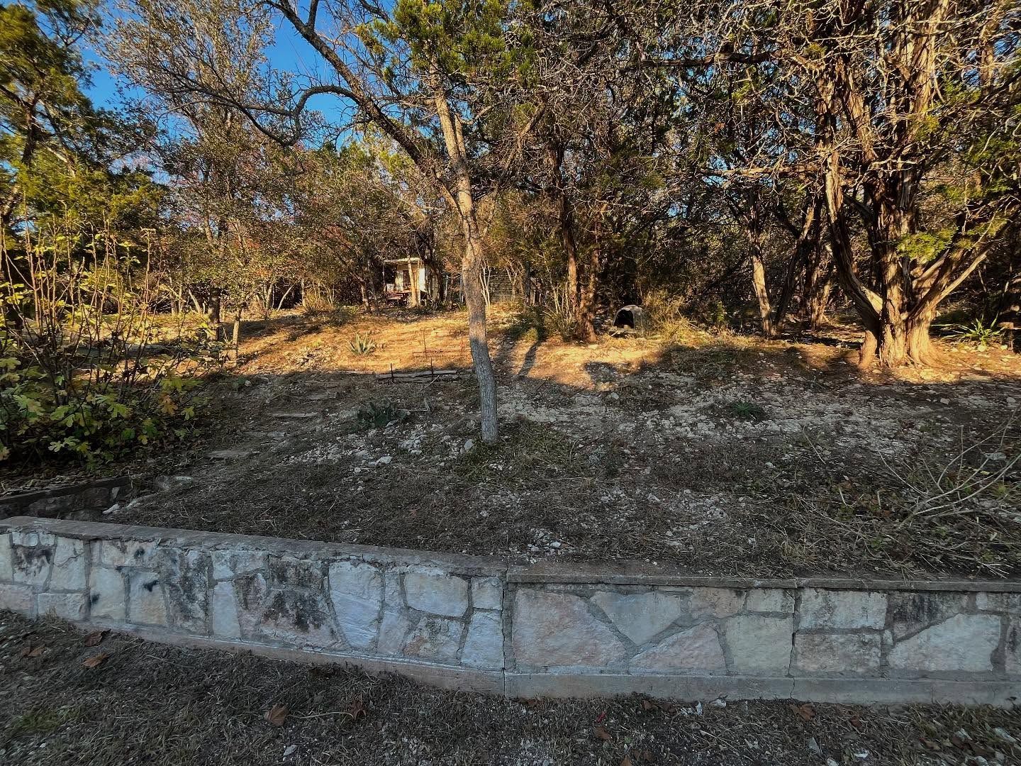 A low stone retaining wall borders a sloped, rocky yard shaded by mature trees under a clear blue sky.