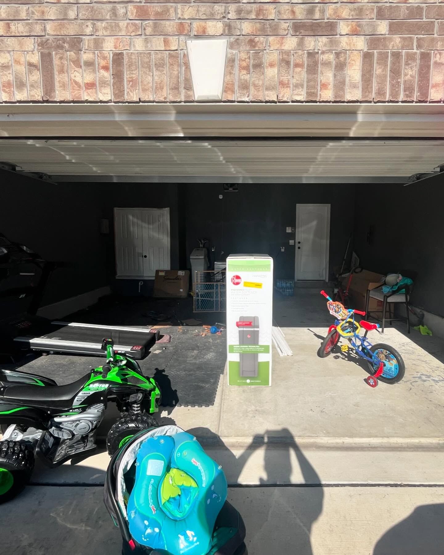 A suburban garage filled with a toy ATV, a bicycle, a box for a tall fan, and various household items.