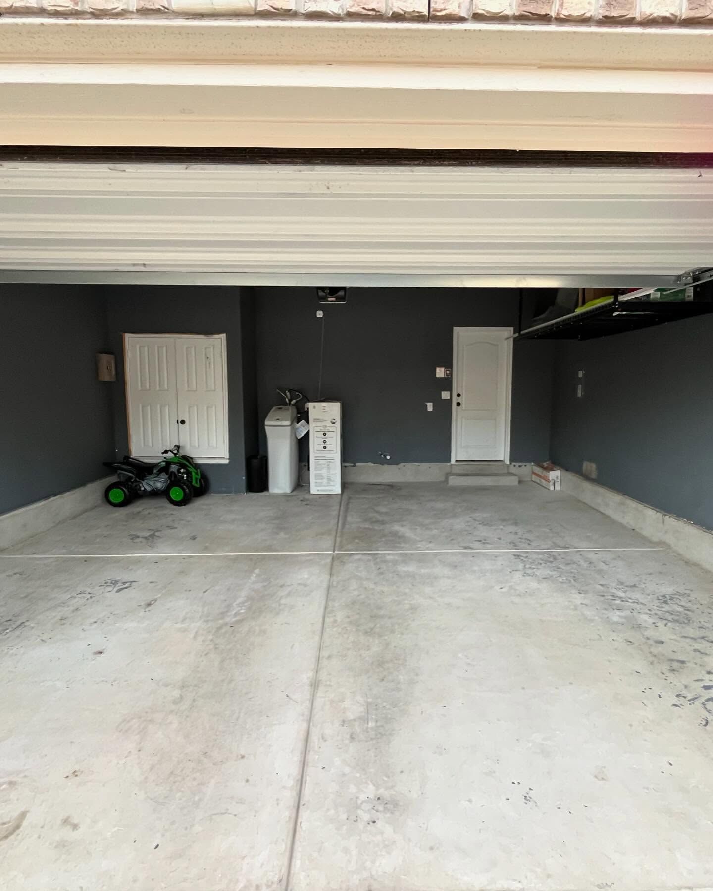 A clean, empty two-car garage with gray walls, a concrete floor, a storage unit, a water softener, and a toy ATV.