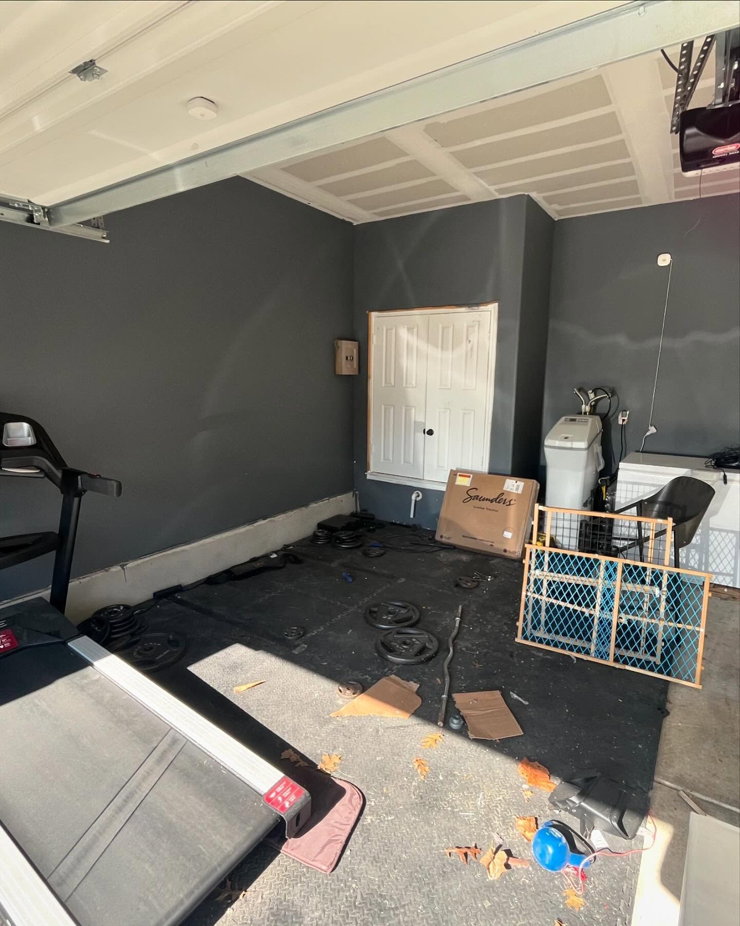 A garage home gym with a treadmill, rubber floor mats, weight plates, a box, and a playpen on a dark gray concrete floor.