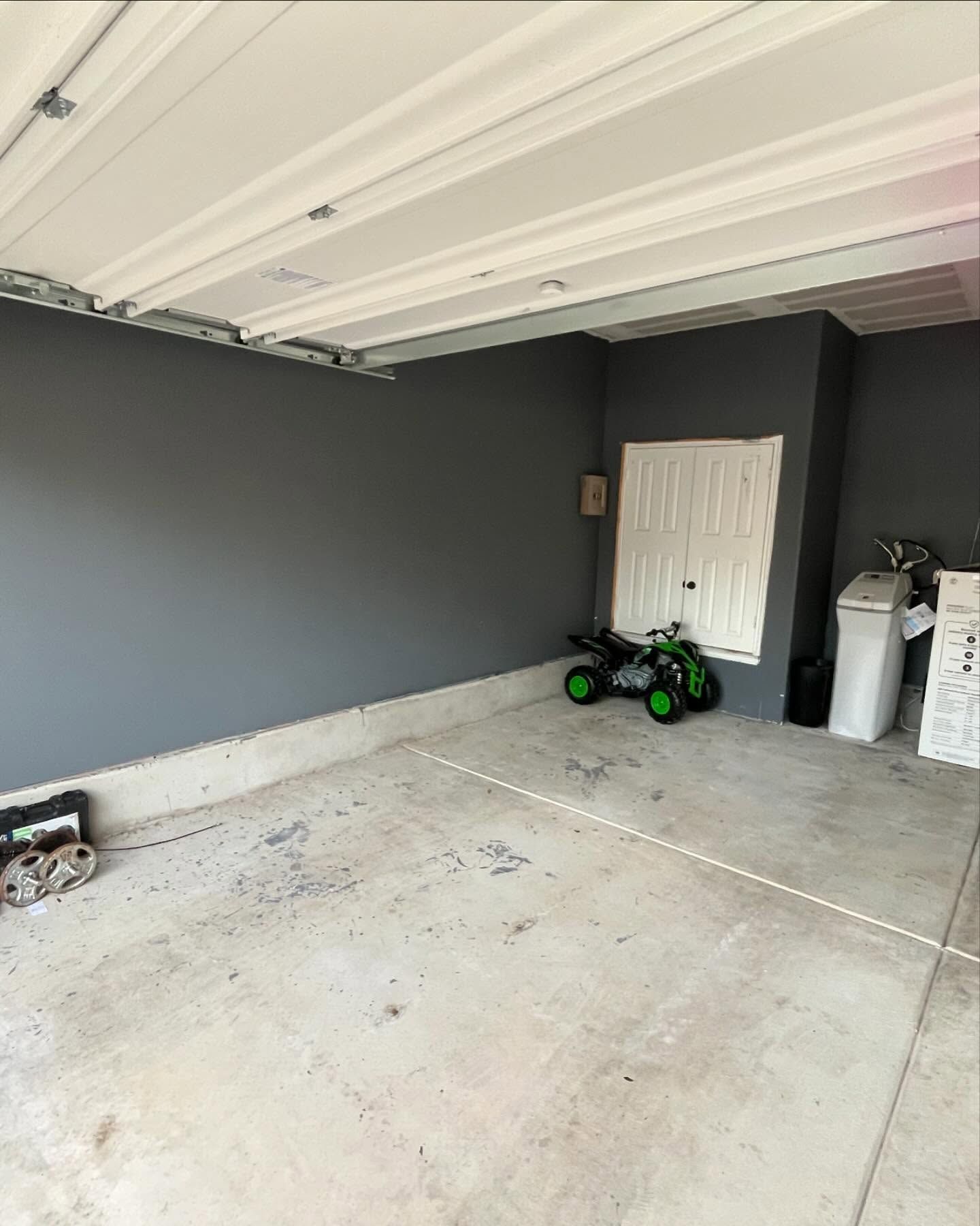 A gray-walled residential garage with a concrete floor, a small green toy ATV, a white storage closet, and a white bin.