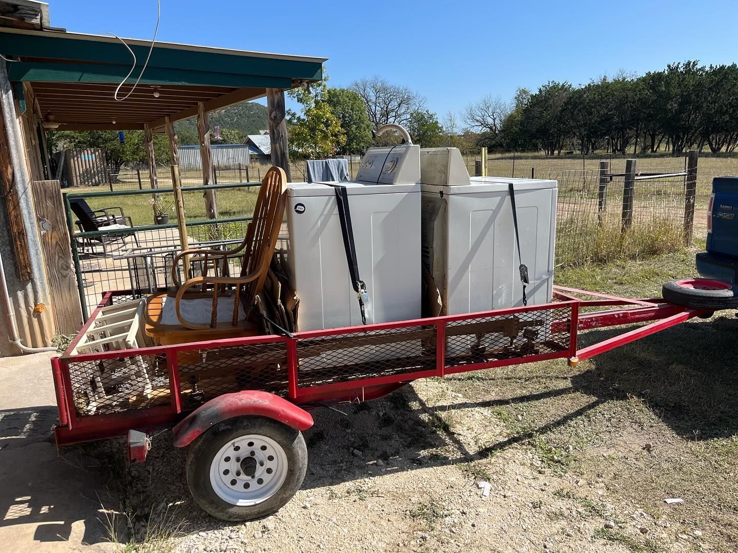 A red utility trailer parked outdoors holds a wicker chair and two white appliances, secured with black straps.