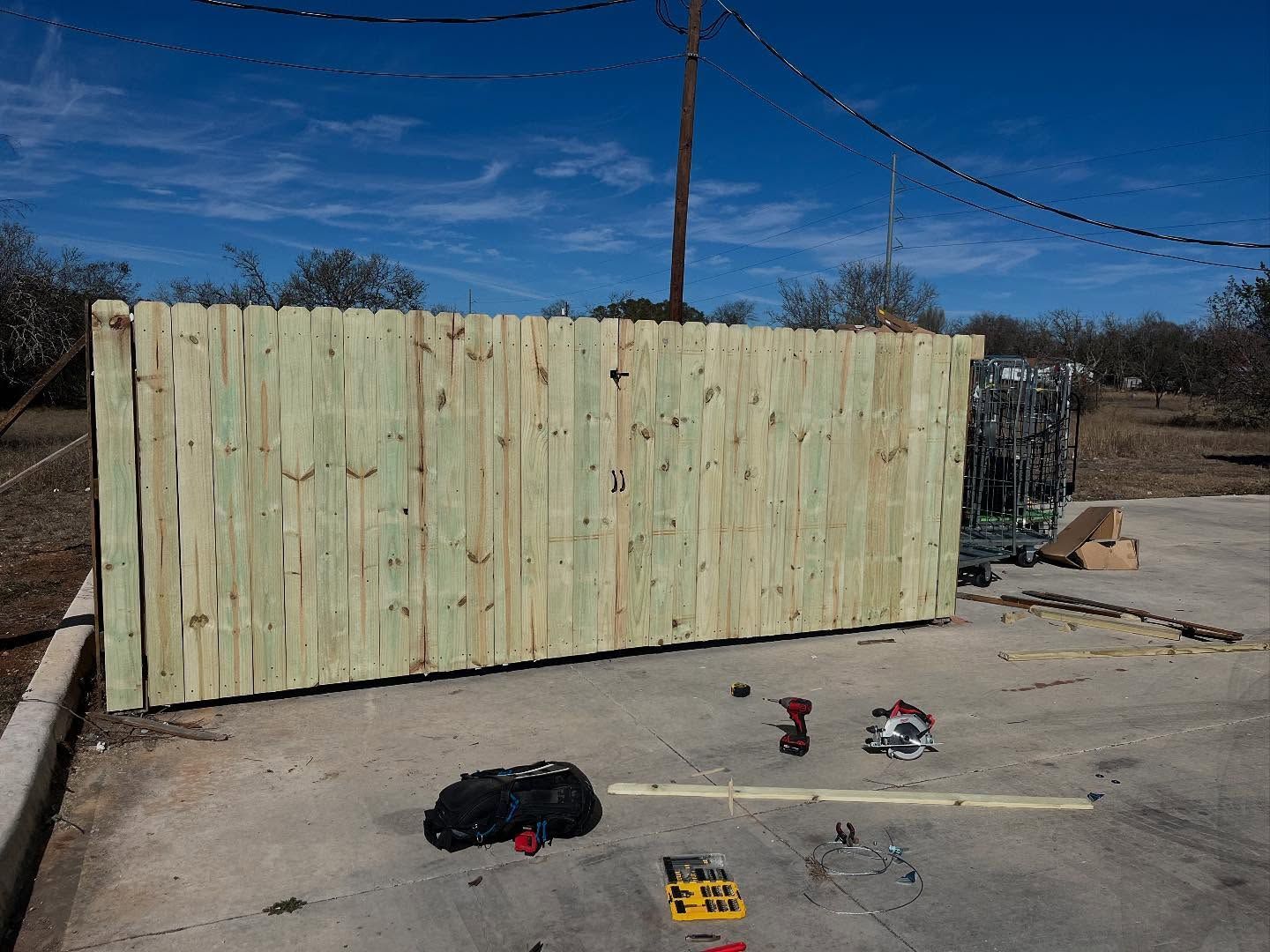 A newly constructed wooden fence section stands on a concrete surface, with power tools lying on the ground nearby.