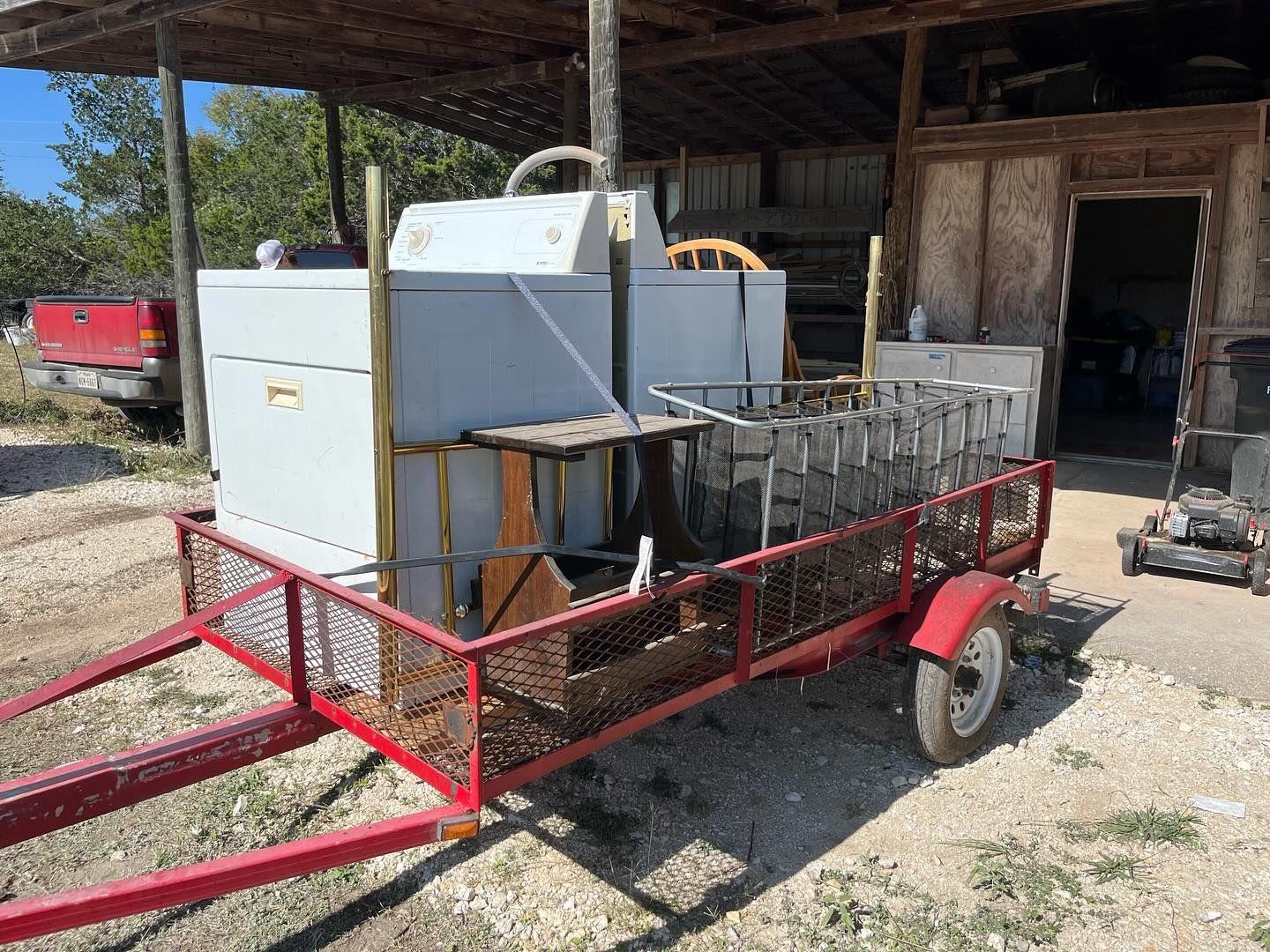 A red utility trailer parked outdoors holds a white appliance, a wooden chair, and a metal wire basket.