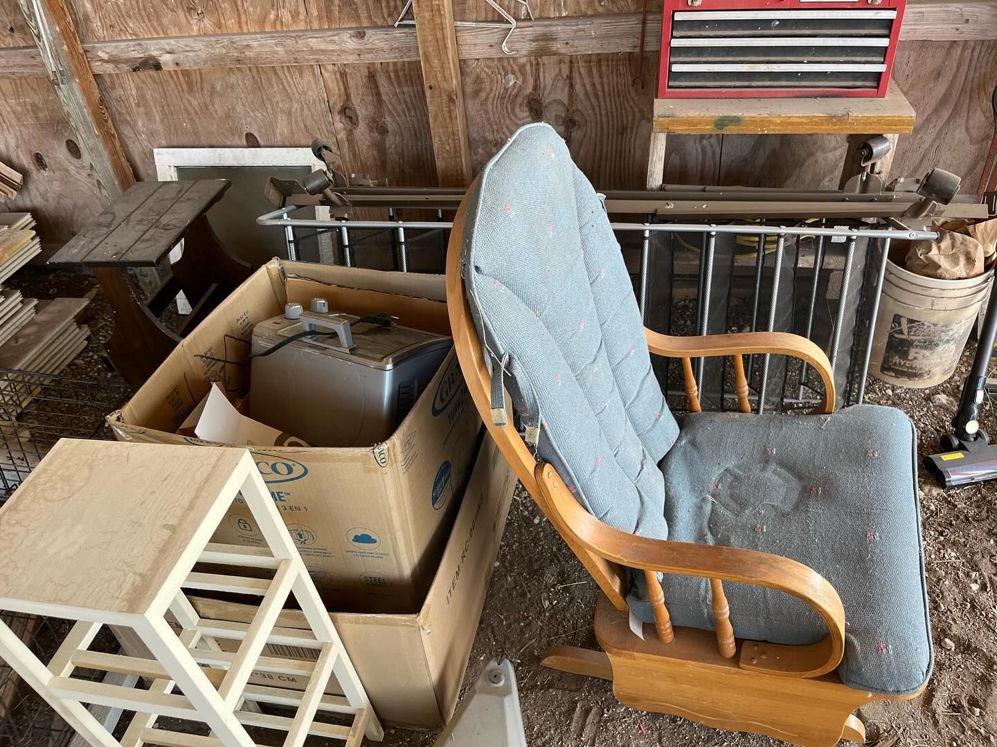 A wooden rocking chair with blue cushions sits beside cardboard boxes and miscellaneous items in a rustic shed.