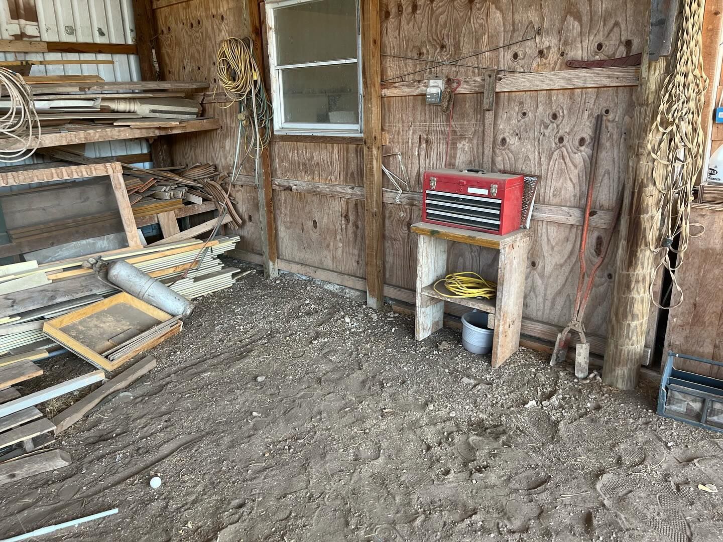 A cluttered barn interior with a small wooden shelf, a red tool chest, a window, and wood scraps scattered on a dirt floor.