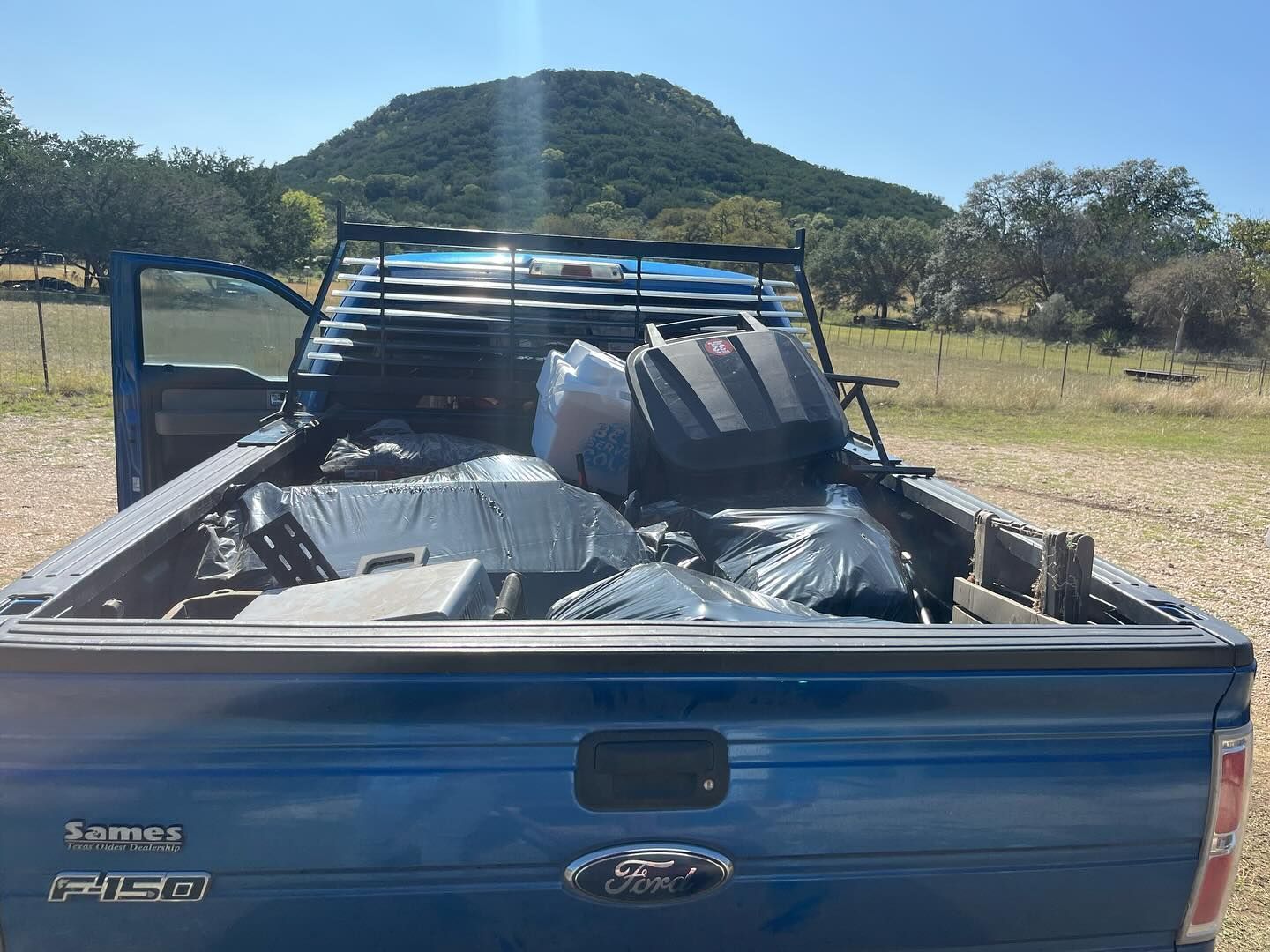 A blue Ford F-150 pickup truck parked in a rural, sunny field, with its bed filled with various items and black bags.