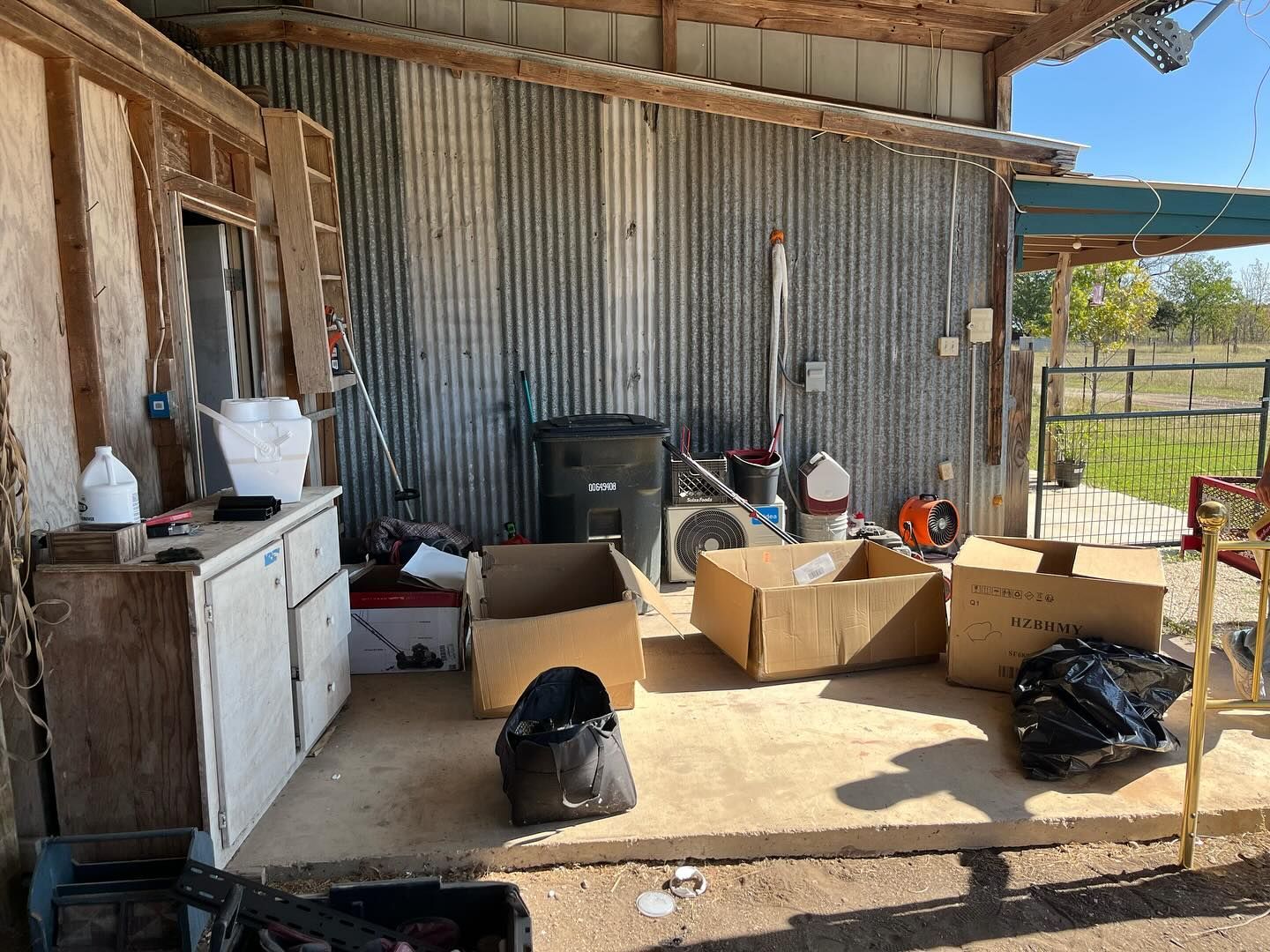 A covered outdoor porch with a wooden cabinet, several cardboard boxes, a trash bin, and bags on a concrete floor.
