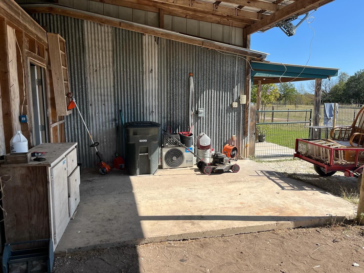 A covered outdoor area with a white cabinet, a black trash bin, power equipment, and a red trailer on a concrete slab.