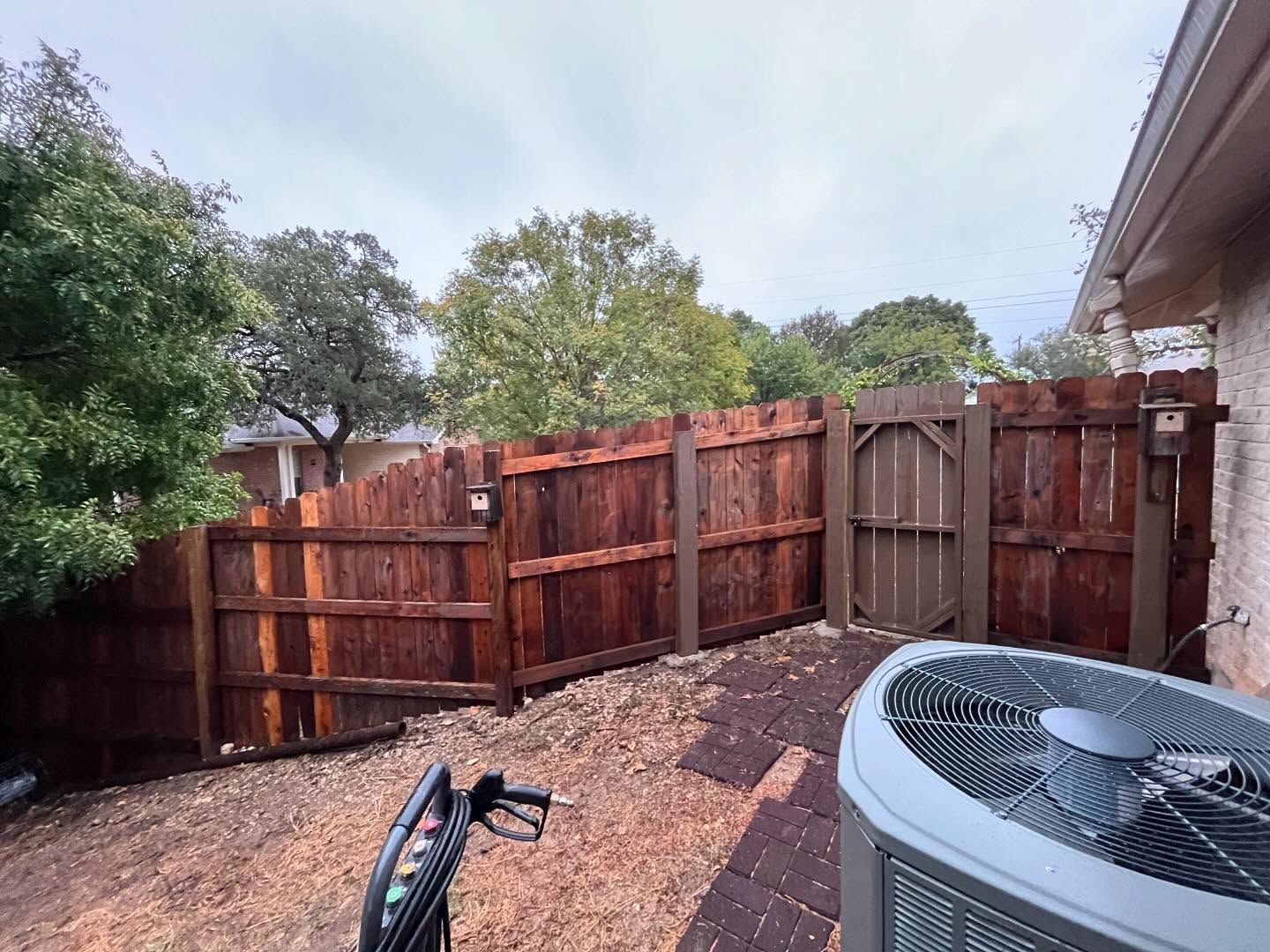 A wooden privacy fence borders a backyard with mulch, trees, and an air conditioning unit.