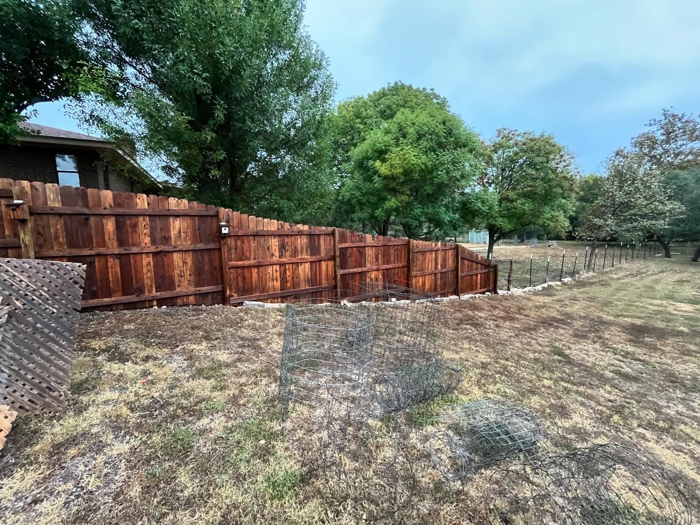 A dark wooden privacy fence runs along a grassy yard under a cloudy, overcast sky.