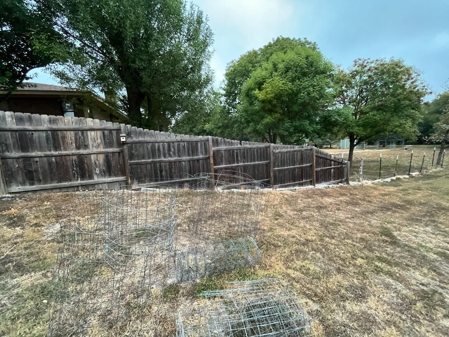 A stepped wooden fence separates a dry grassy yard from trees and an open field under a cloudy sky.
