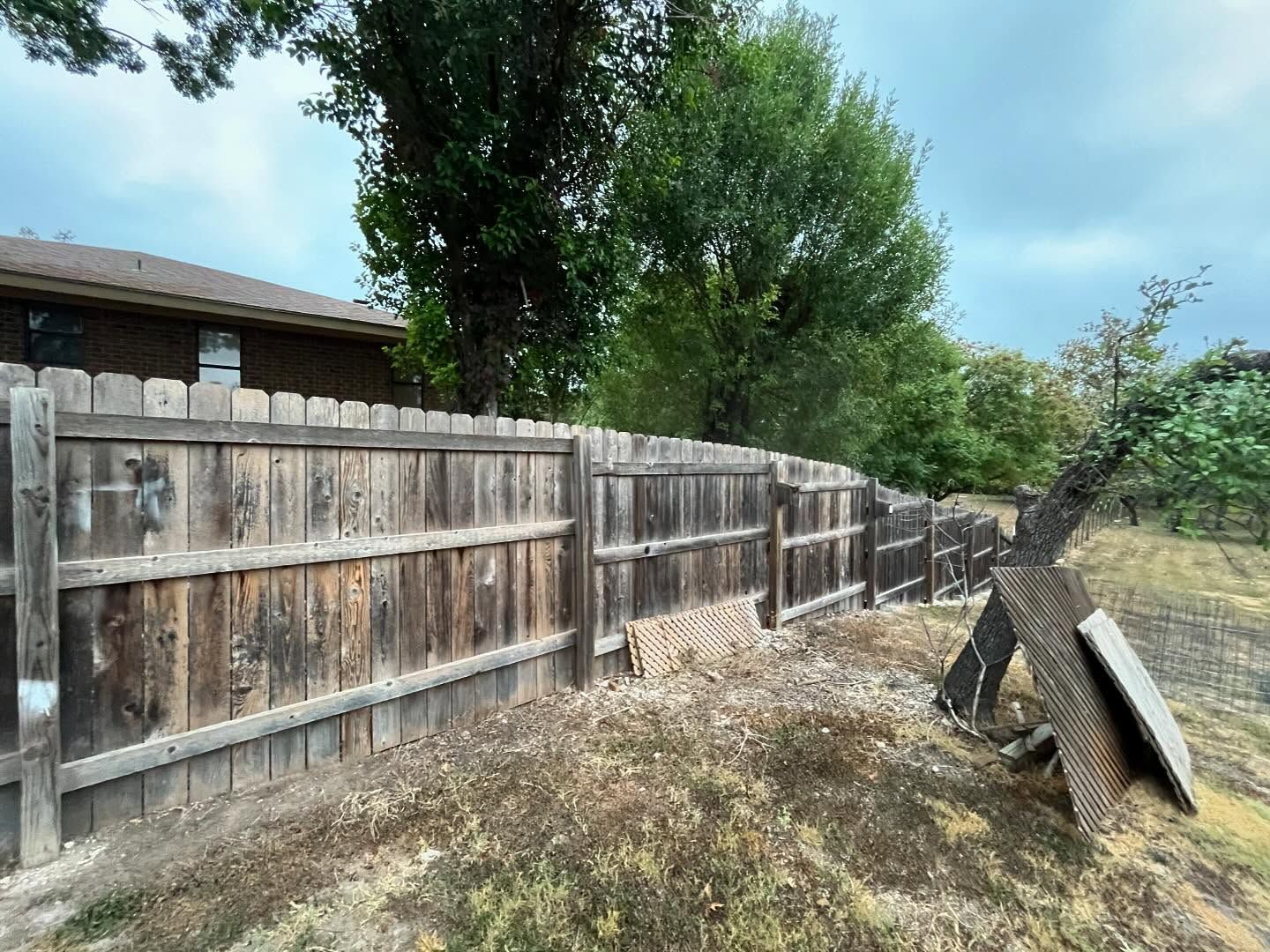 A weathered wooden privacy fence runs along a yard edge next to a brick house, with trees and a fallen fence post nearby.